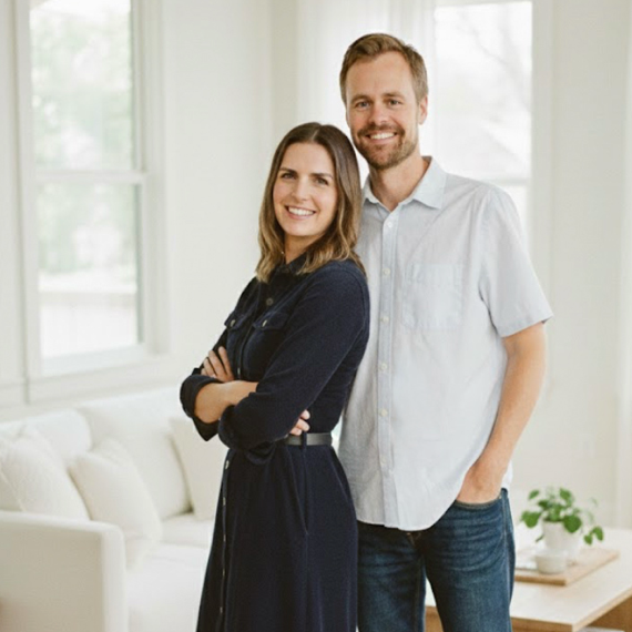 A smiling woman with crossed arms and a man standing beside her, both indoors in a well-lit room with large windows and a white couch.