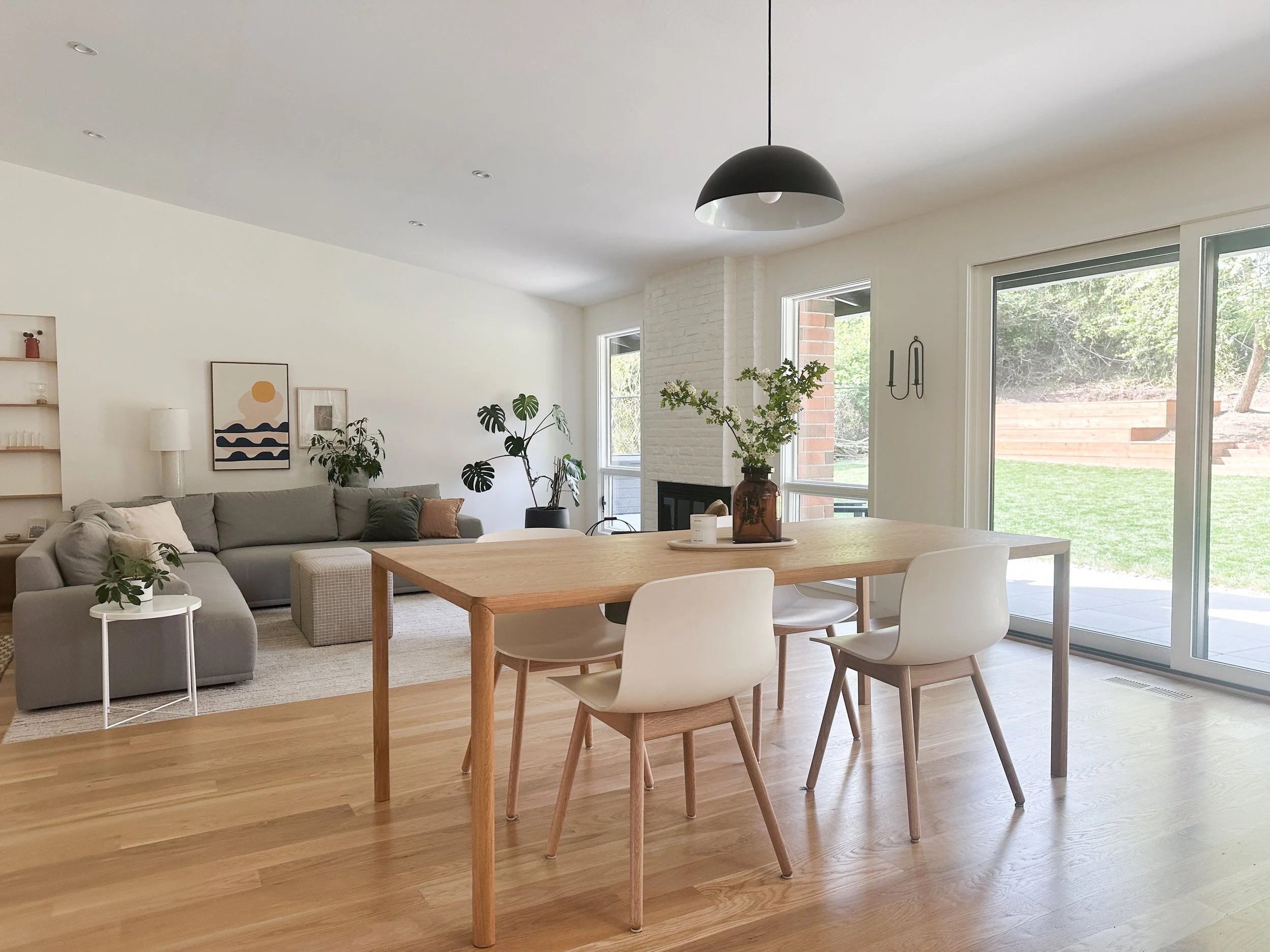 Bright open living and dining area with a wooden dining table, white chairs, a gray sofa, potted plants, artwork on the wall, a fireplace, and large glass doors leading to a backyard.