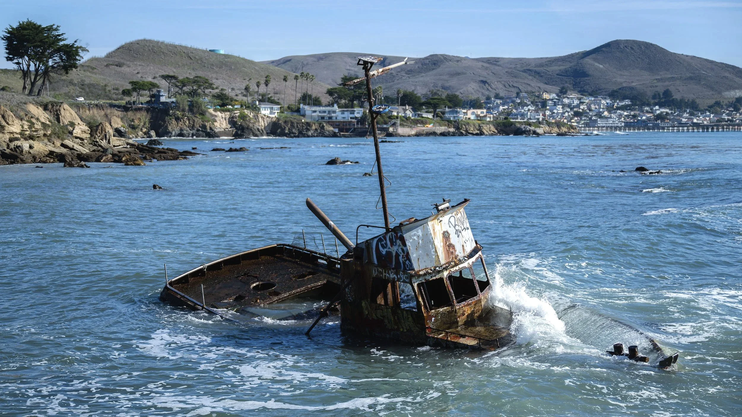 A rusted, sunken boat partially submerged in the ocean near a rocky shoreline with houses and hills in the background. Shell Beach, California