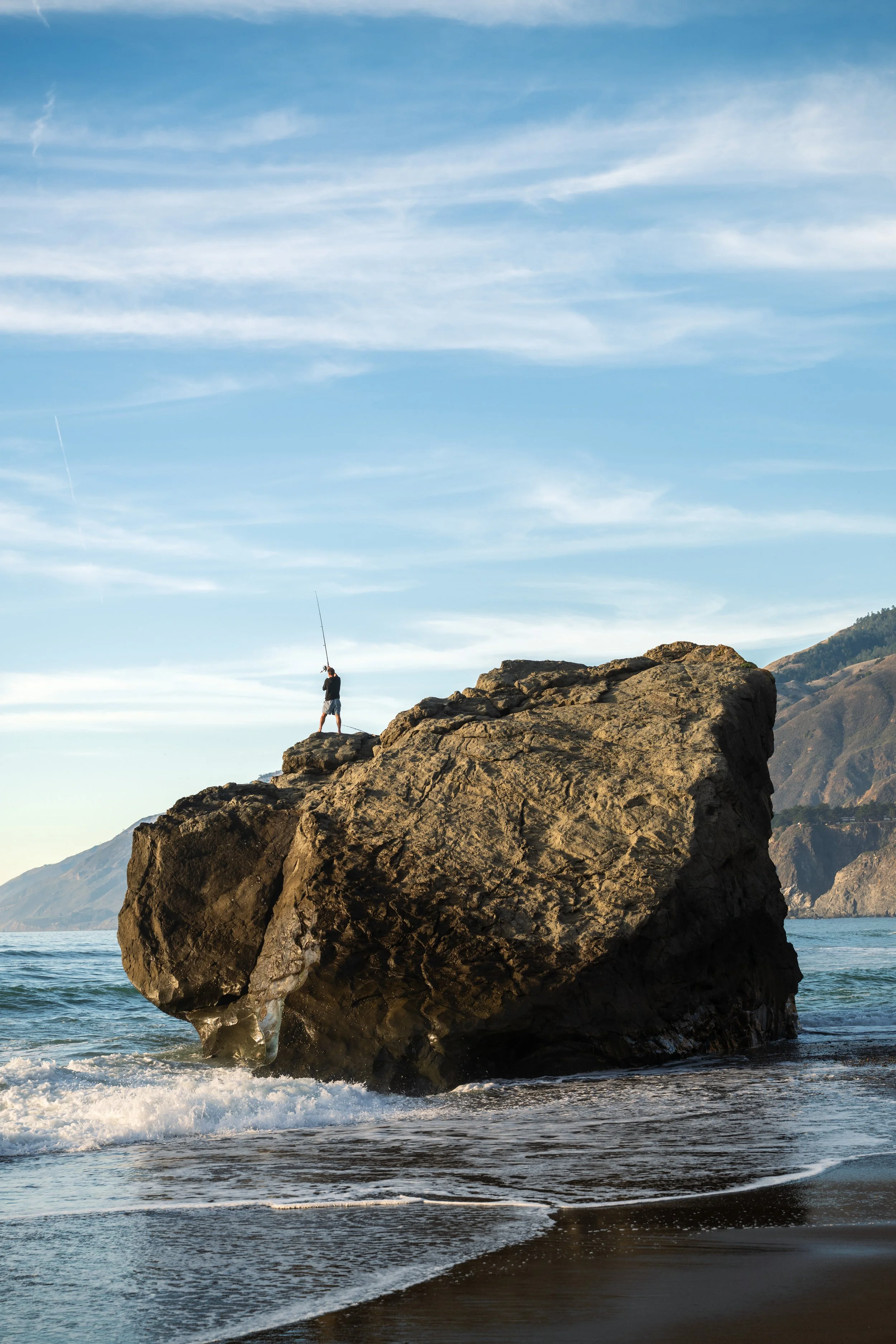 Person fishing on a large rock by the ocean with mountains in the background and a partly cloudy sky. Big Sur, California