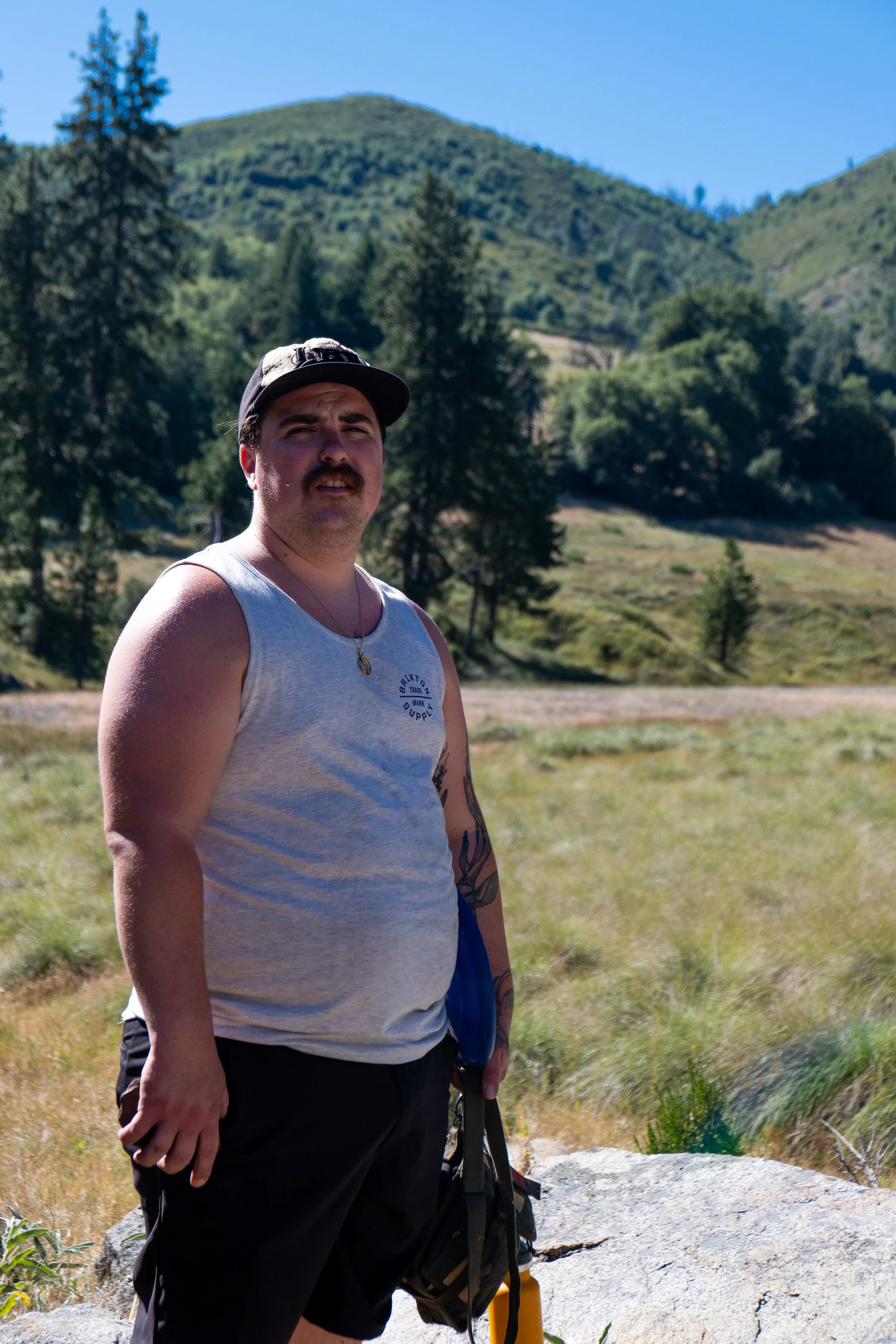A man with a mustache and dark hair standing outdoors in front of a mountainous landscape with trees and grassy terrain, wearing a gray sleeveless shirt, black shorts, a cap, and holding a backpack. Palomar Mountain, California