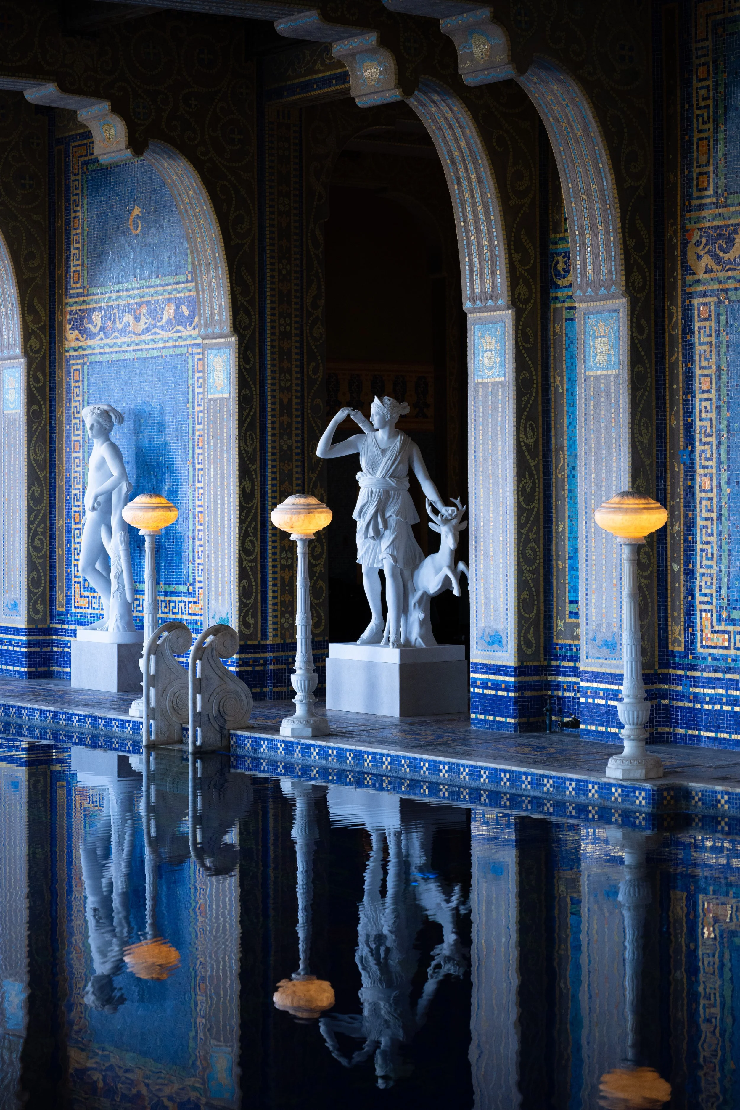 Interior of a luxurious building with blue tiled walls, white marble statues of women, illuminated by tall, elegant lamps, and reflected in a water pool in the foreground. Hearst Castle, California