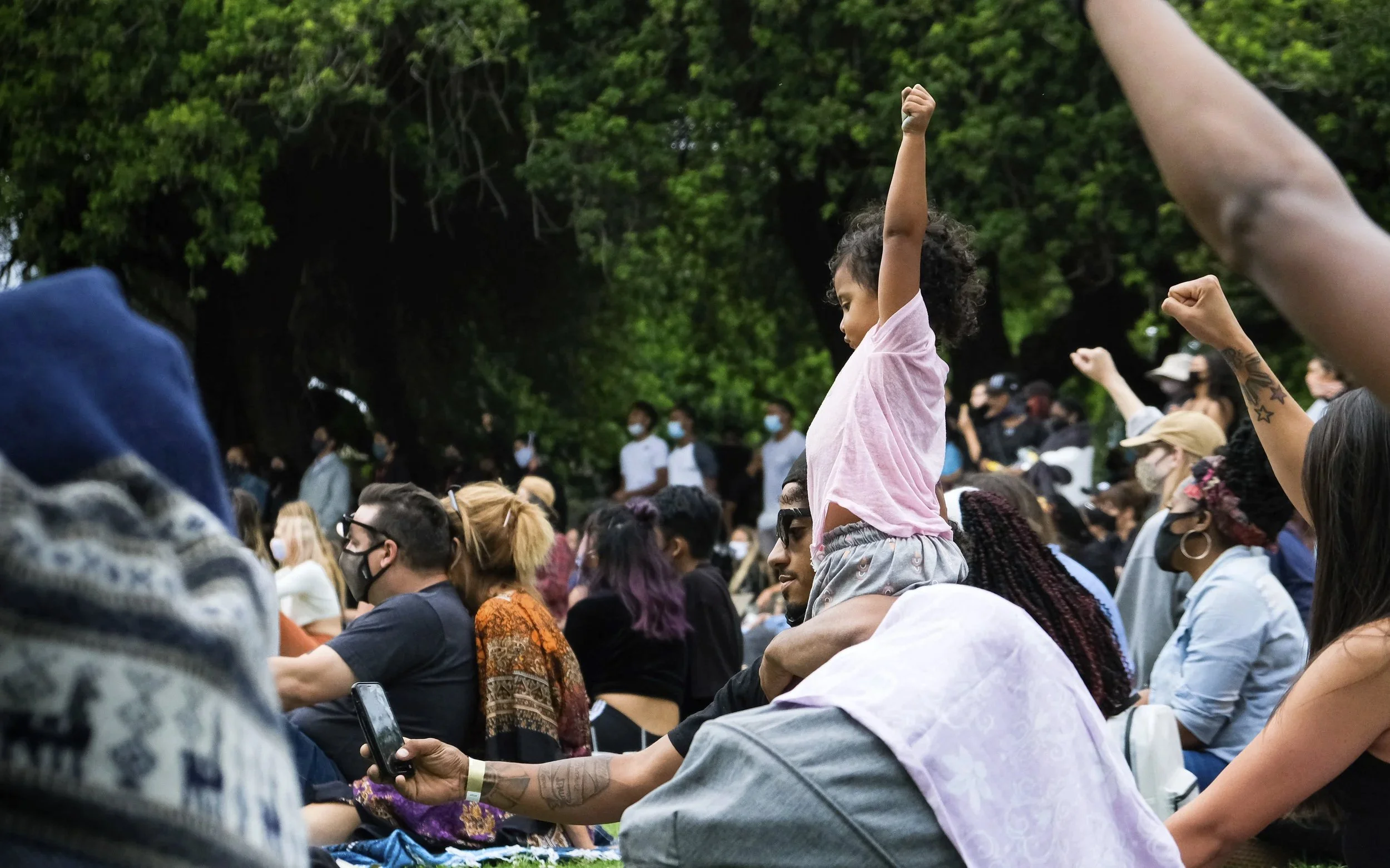 Crowd of diverse people attending a protest or rally outdoors, some raising fists and showing various expressions, with trees in the background. San Diego, California
