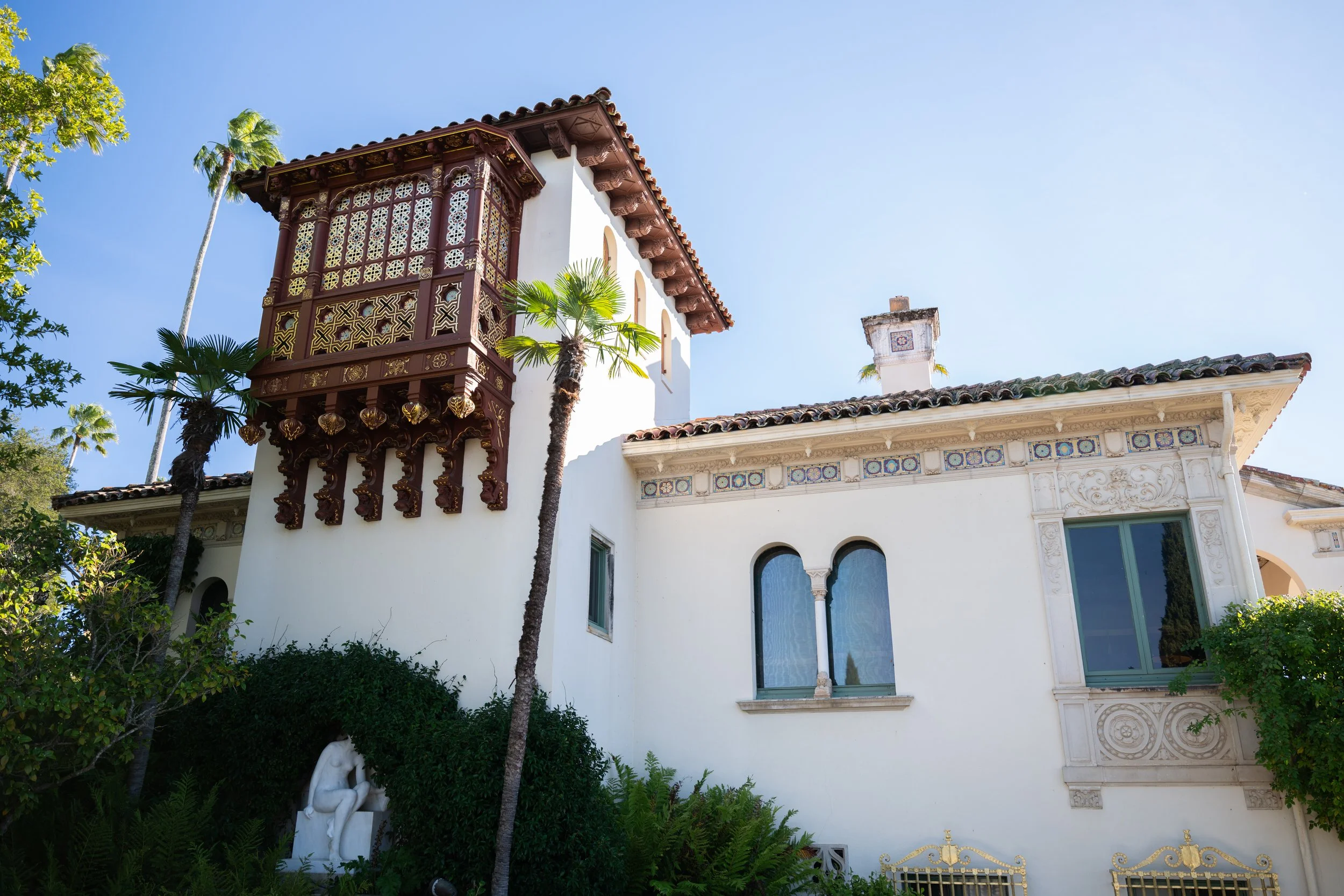 A historic mansion with ornate woodwork and white stucco walls, surrounded by palm trees and greenery. Hearst Castle, California