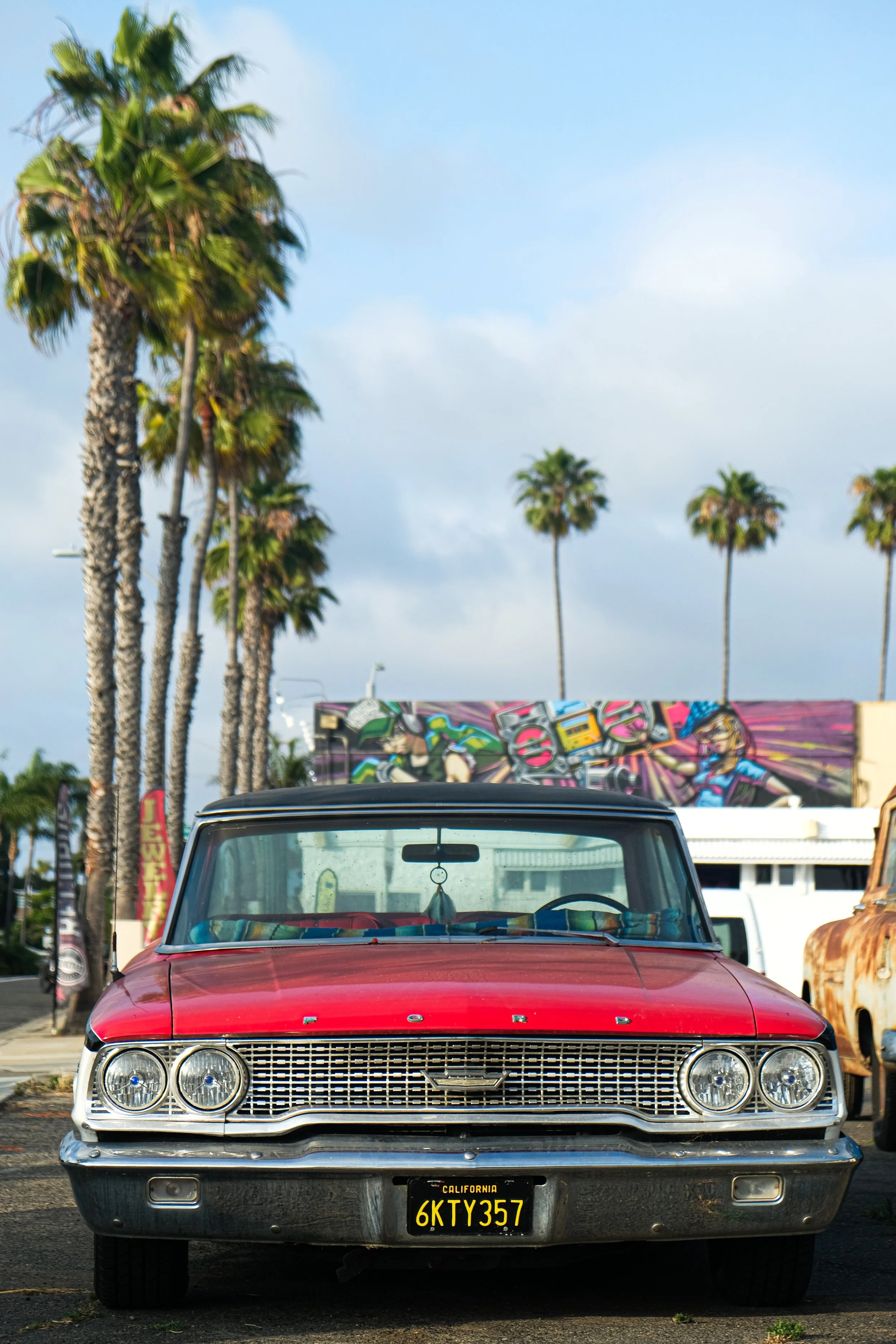 Front view of a vintage red Ford car parked outdoors with palm trees and colorful graffiti wall in the background. Ocean Beach, California