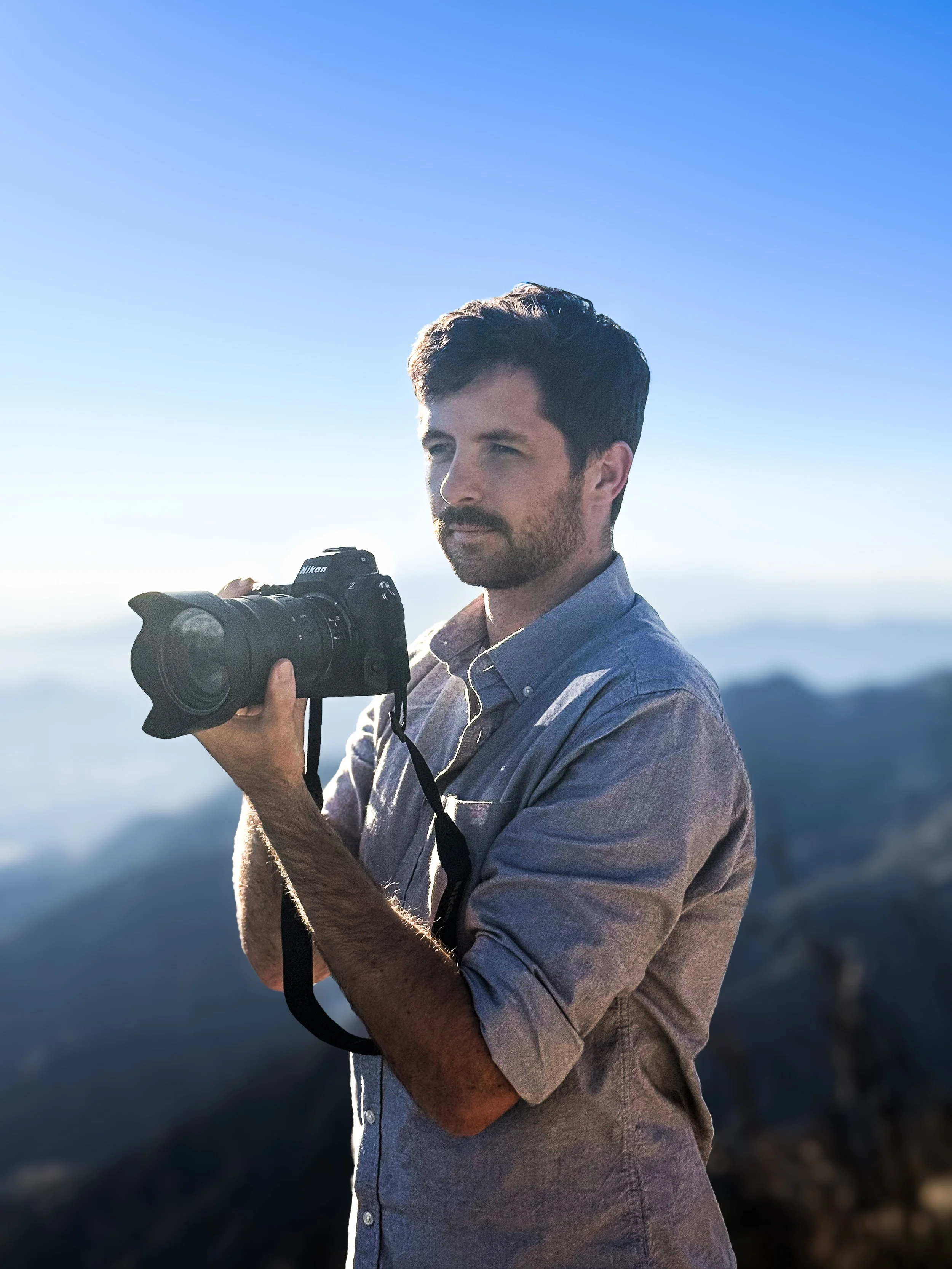 Man with short dark hair and beard holding a Nikon camera outdoors with mountain scenery in the background. Angeles National Forest, California