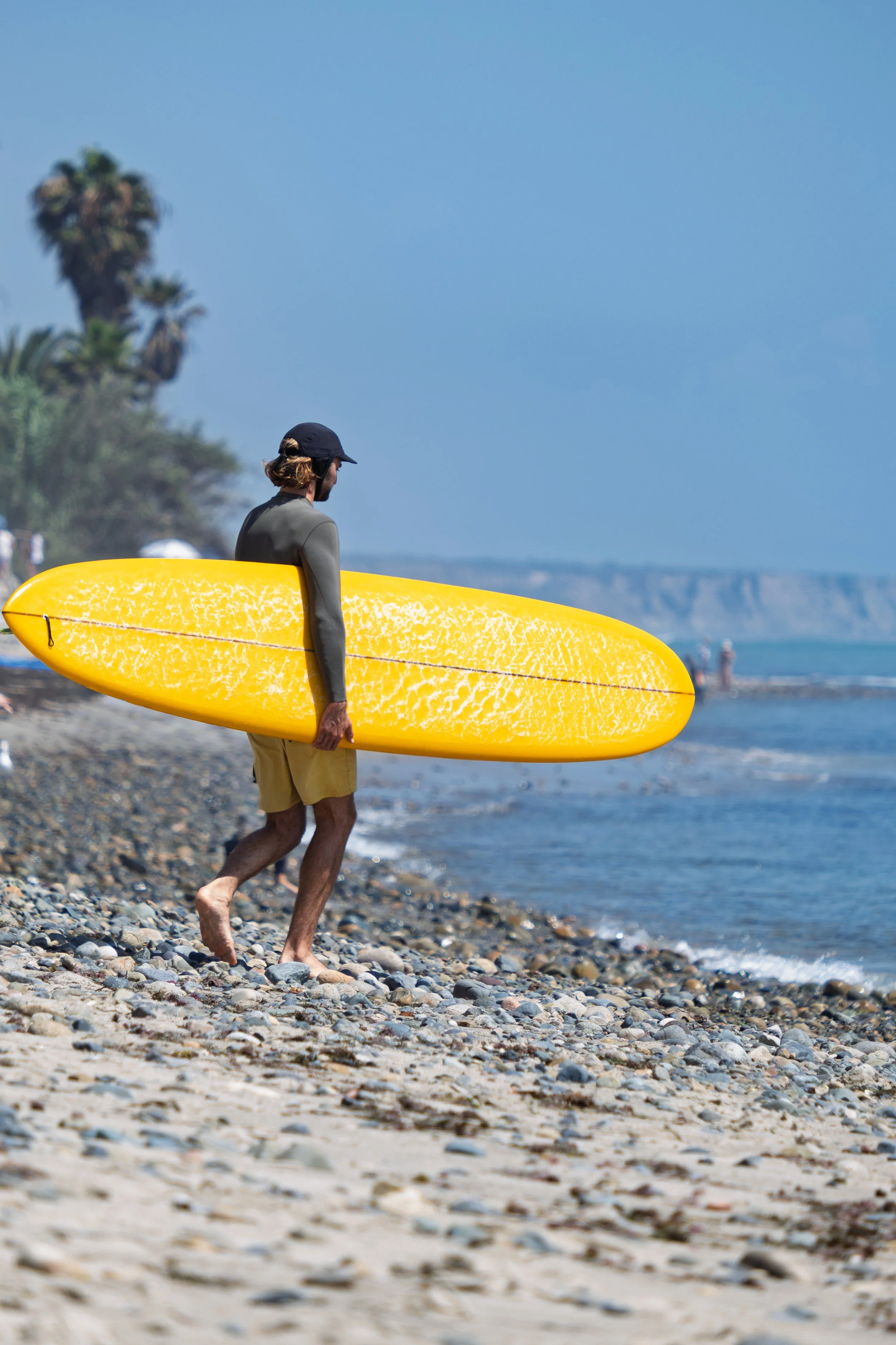 A man walking barefoot on a rocky beach carrying a yellow surfboard, with palm trees and cliffs in the background under a blue sky. San Onofre Beach, California