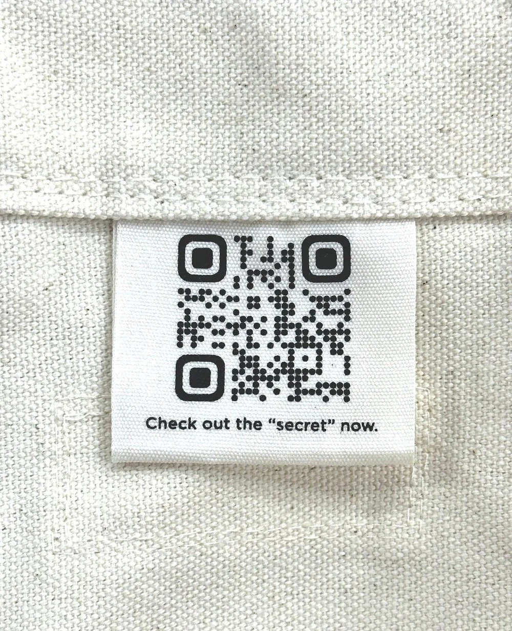 Clothing label with a QR code and the text "Check out the 'secret' now" on a beige fabric background.