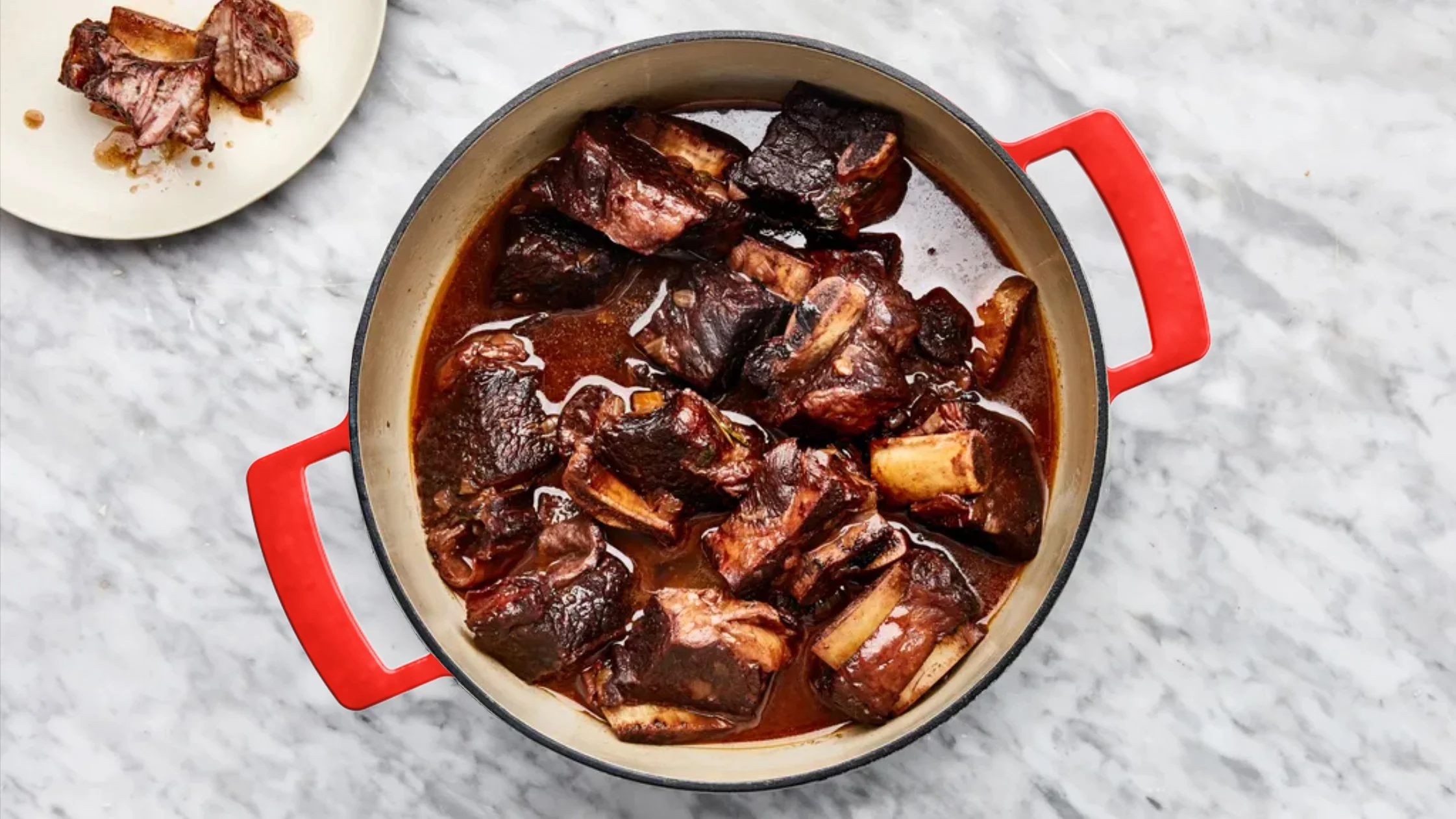 A pot with cooked beef short ribs in a rich, dark sauce, on a white marble countertop; a small plate with some cooked beef pieces is visible in the background.
