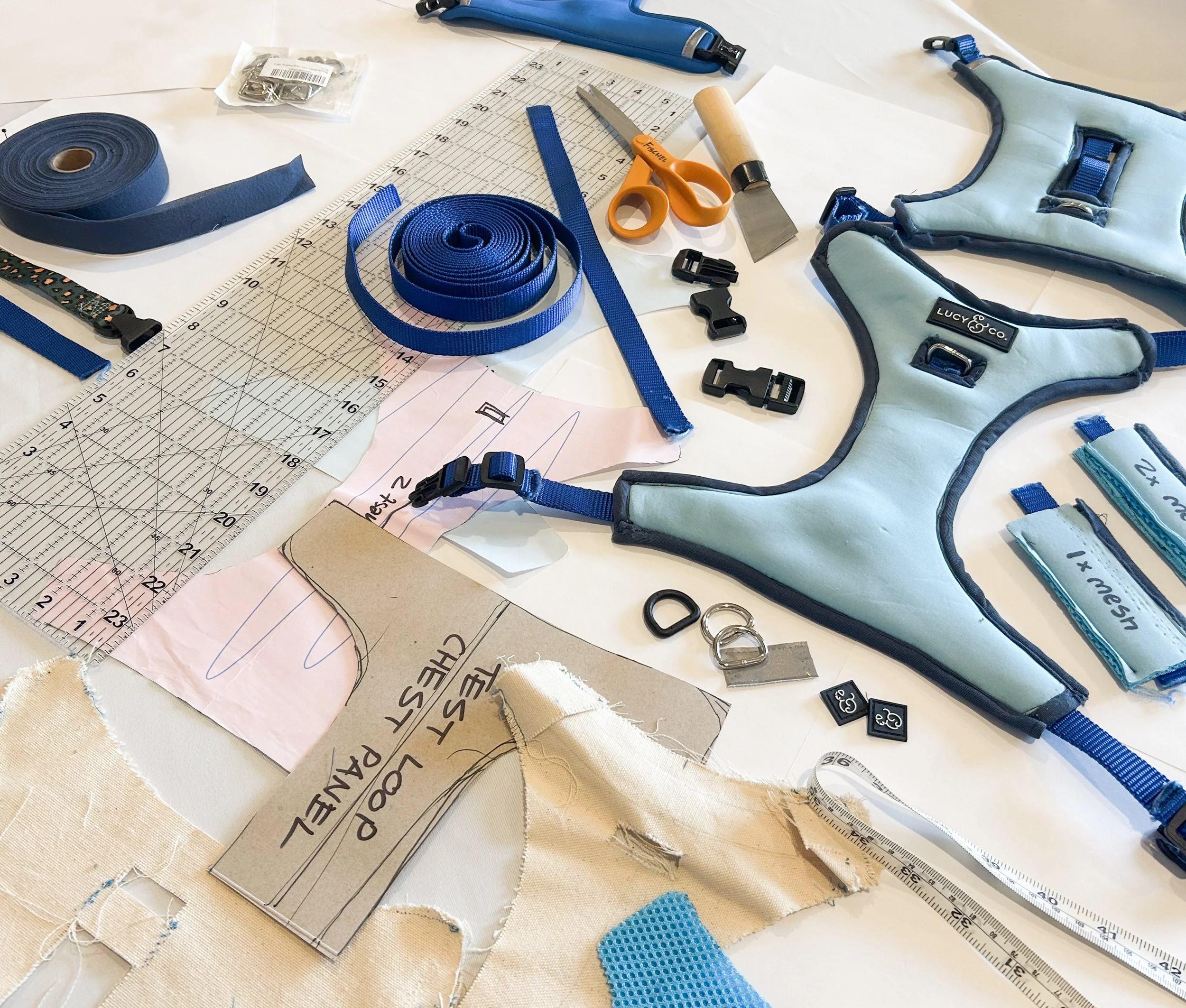 A workspace with fabric, measuring tools, scissors, clips, and a pet harness, indicating a tailoring or sewing project.  Lucy & Co. no-pull harness.