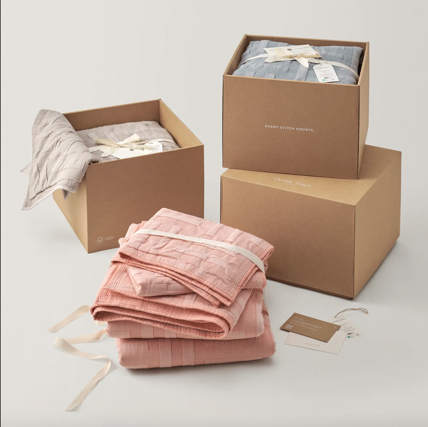 Stack of pink and beige folded blankets with tags, two open cardboard boxes containing folded linens, and a closed box in the background against a light backdrop.
