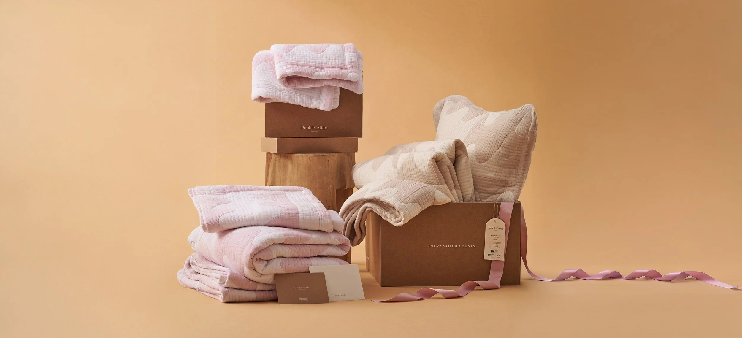 Pink and beige textiles, including blankets and pillows, arranged on boxes and a wooden stand against a beige background, with tags and a pink ribbon.