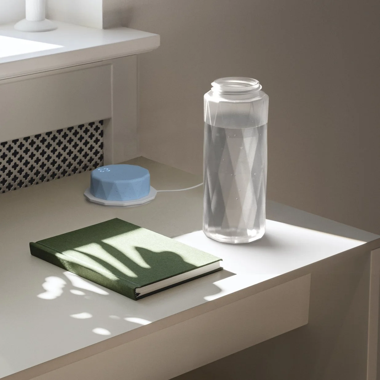 A white desk with a closed green notebook, a large transparent water bottle, and a small blue electric device with a white cord. Sunlight casts shadows on the desk.