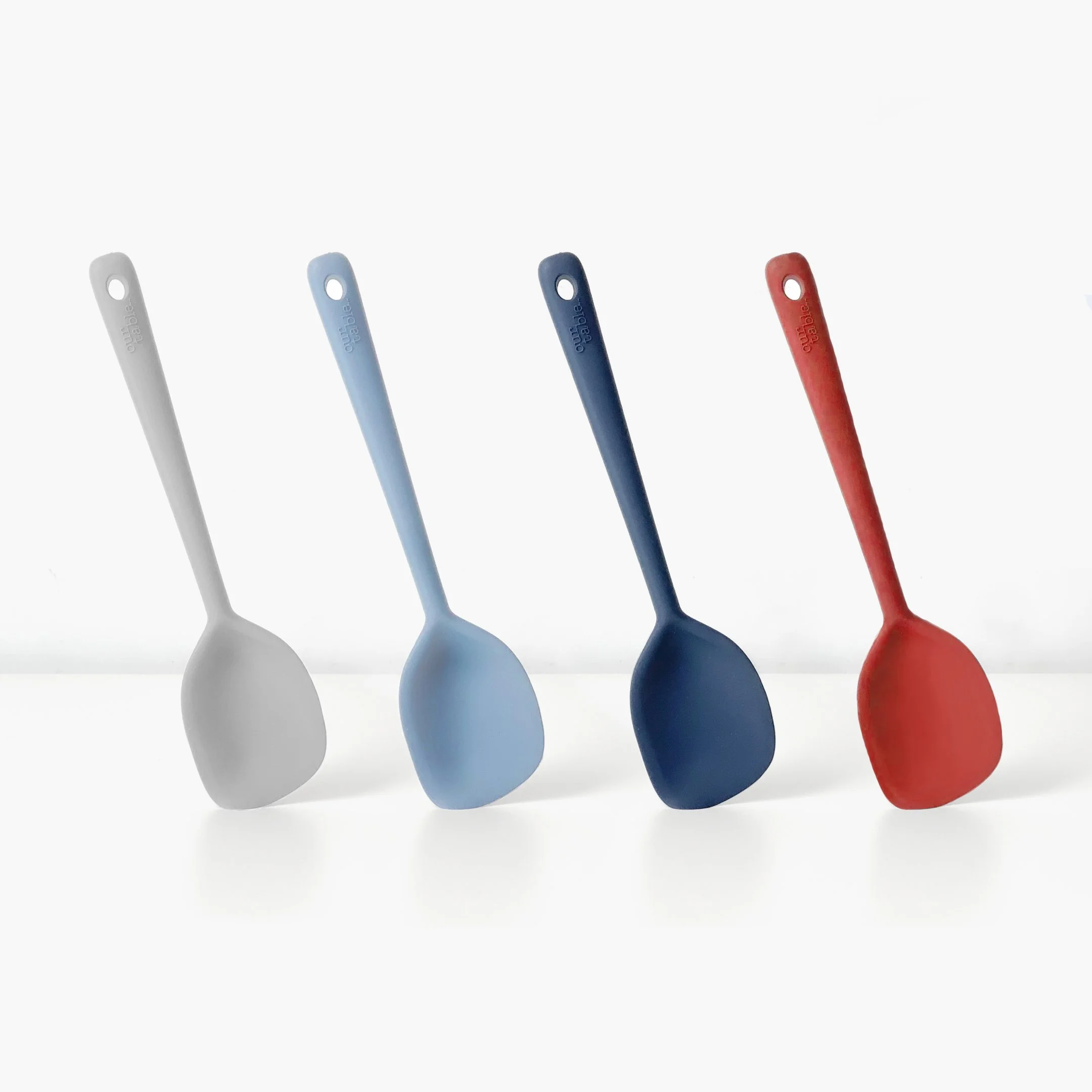 Four colorful silicone spatulas in gray, light blue, dark blue, and red, standing upright on a white surface against a white background.