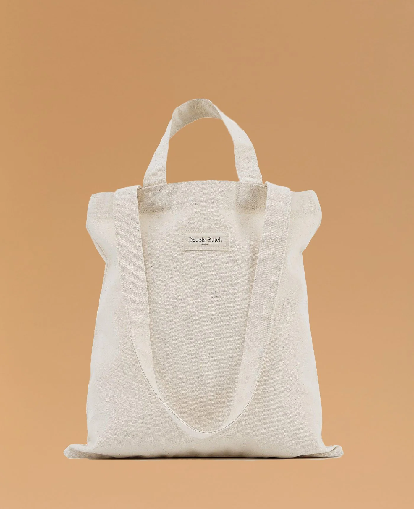 A beige canvas tote bag with shoulder straps and a small zippered pocket, displayed against a plain peach background.