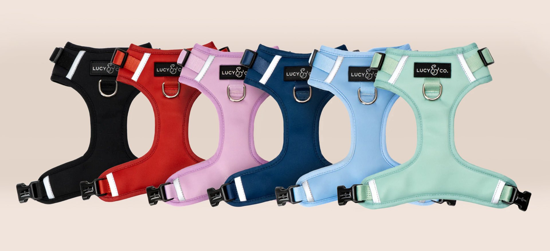 Six colorful dog harnesses in black, red, pink, navy, light blue, and mint green, arranged in a row on a neutral background.  Lucy & Co. no-pull harness.