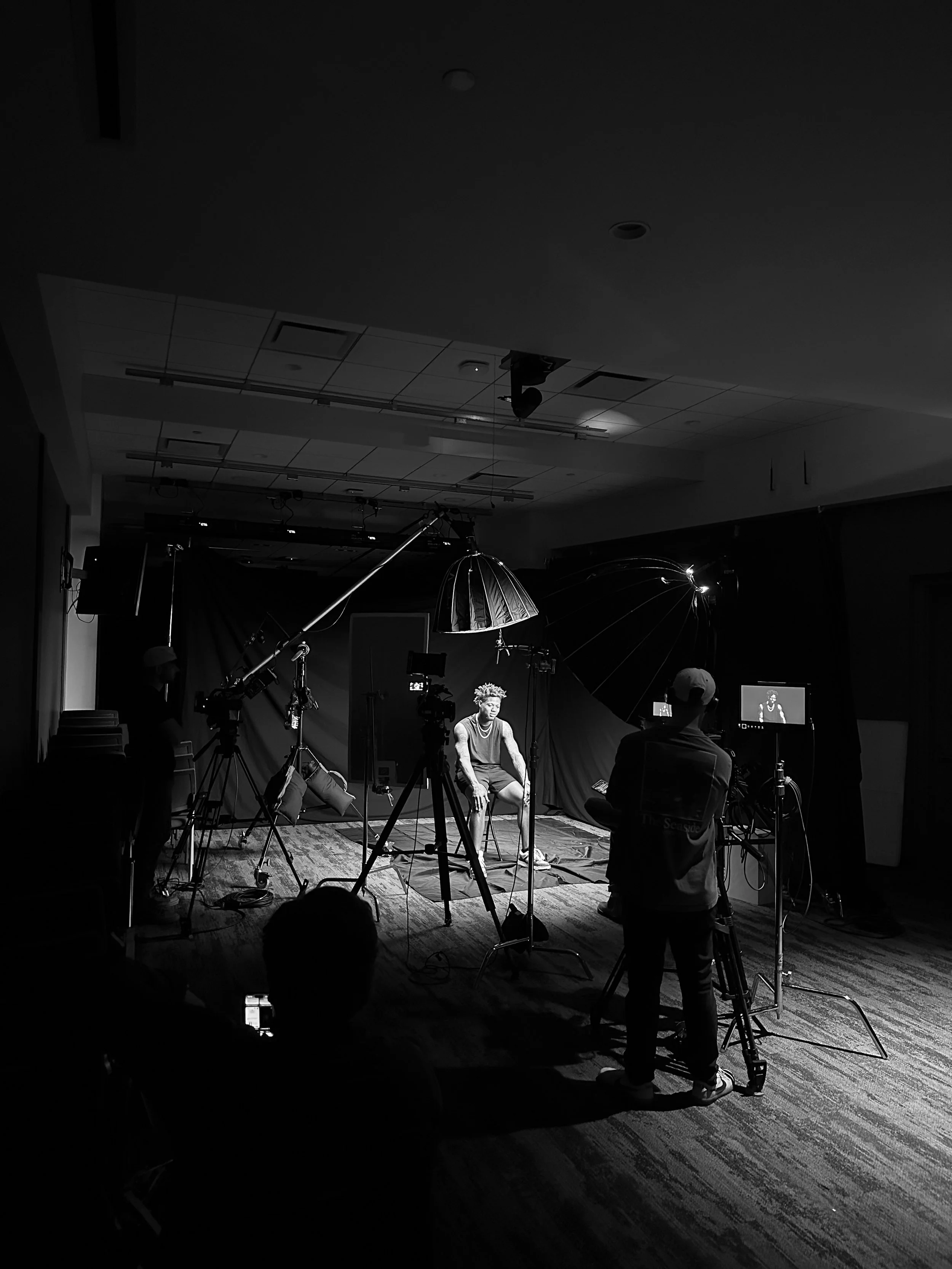 A black and white photograph of a video or photo shoot in a studio, with a person seated in the center surrounded by cameras, lights, and crew members.