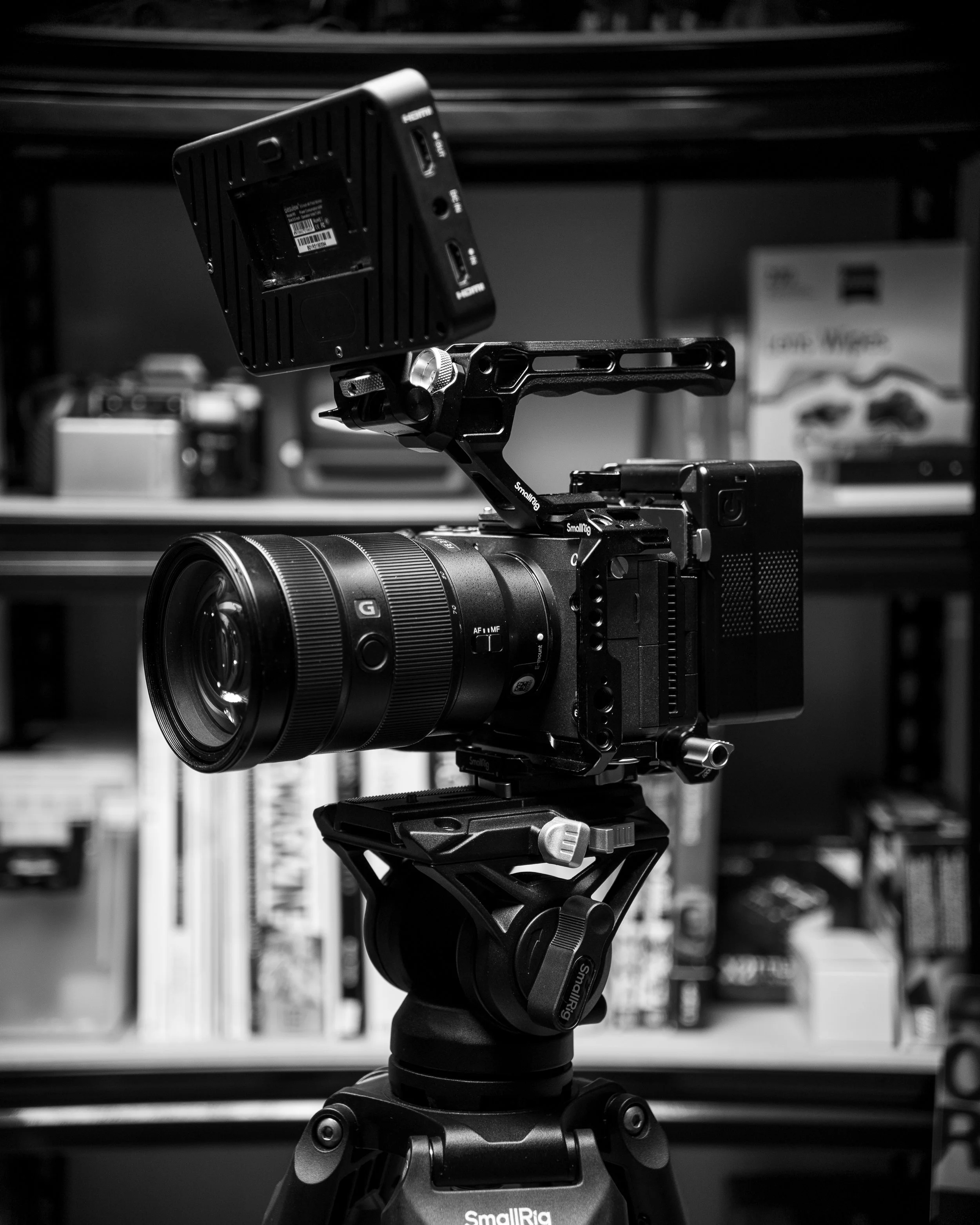 Black and white photo of a professional camera mounted on a tripod, with an external monitor attached on top. Shelves with books, boxes, and camera equipment are visible in the background.
