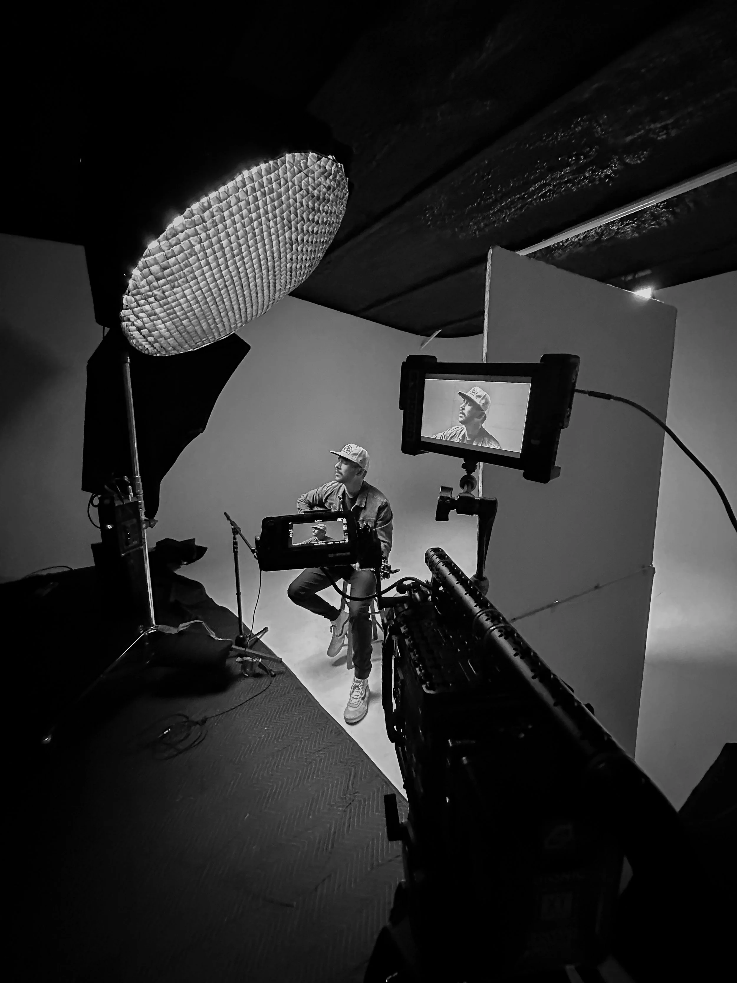 Black and white photo of a man sitting on a stool in a studio, being recorded with professional video equipment, including lights and cameras, with a backdrop behind him.