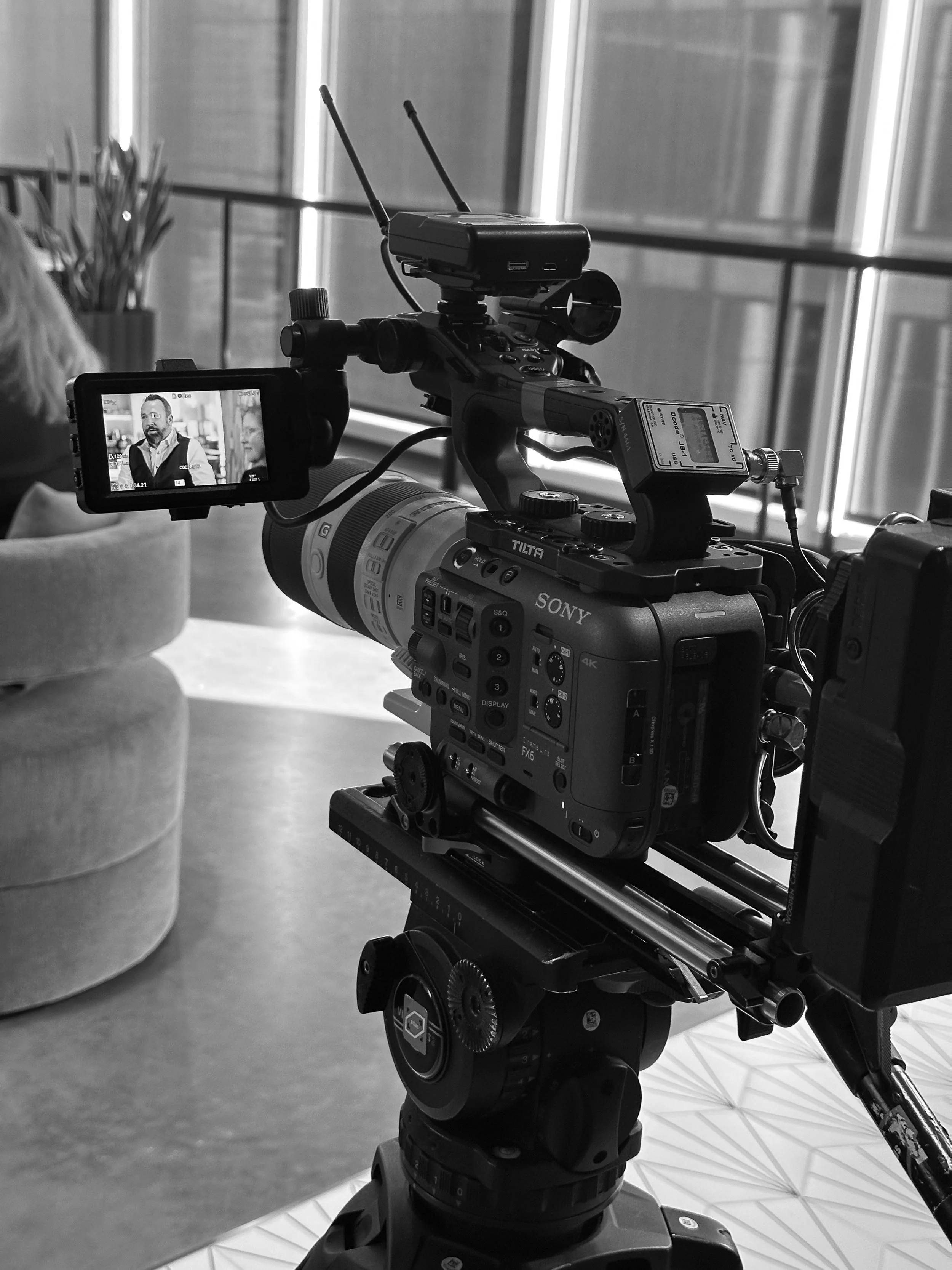 A professional video camera on a tripod indoors, capturing a scene with a person on a monitor display.