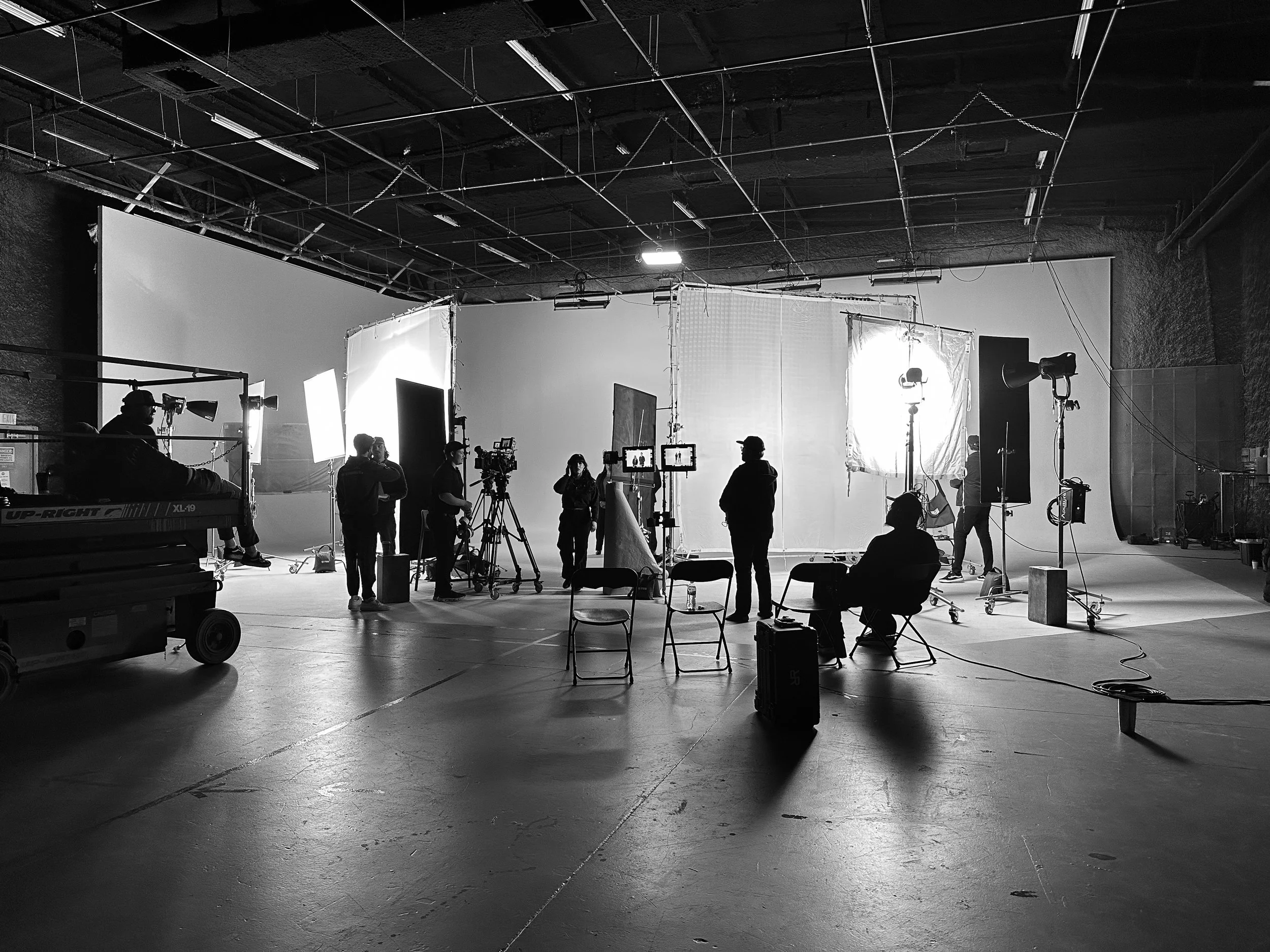 Black and white photo of a film or photo studio with crew members, cameras, lighting equipment, and backdrops preparing for a shoot.