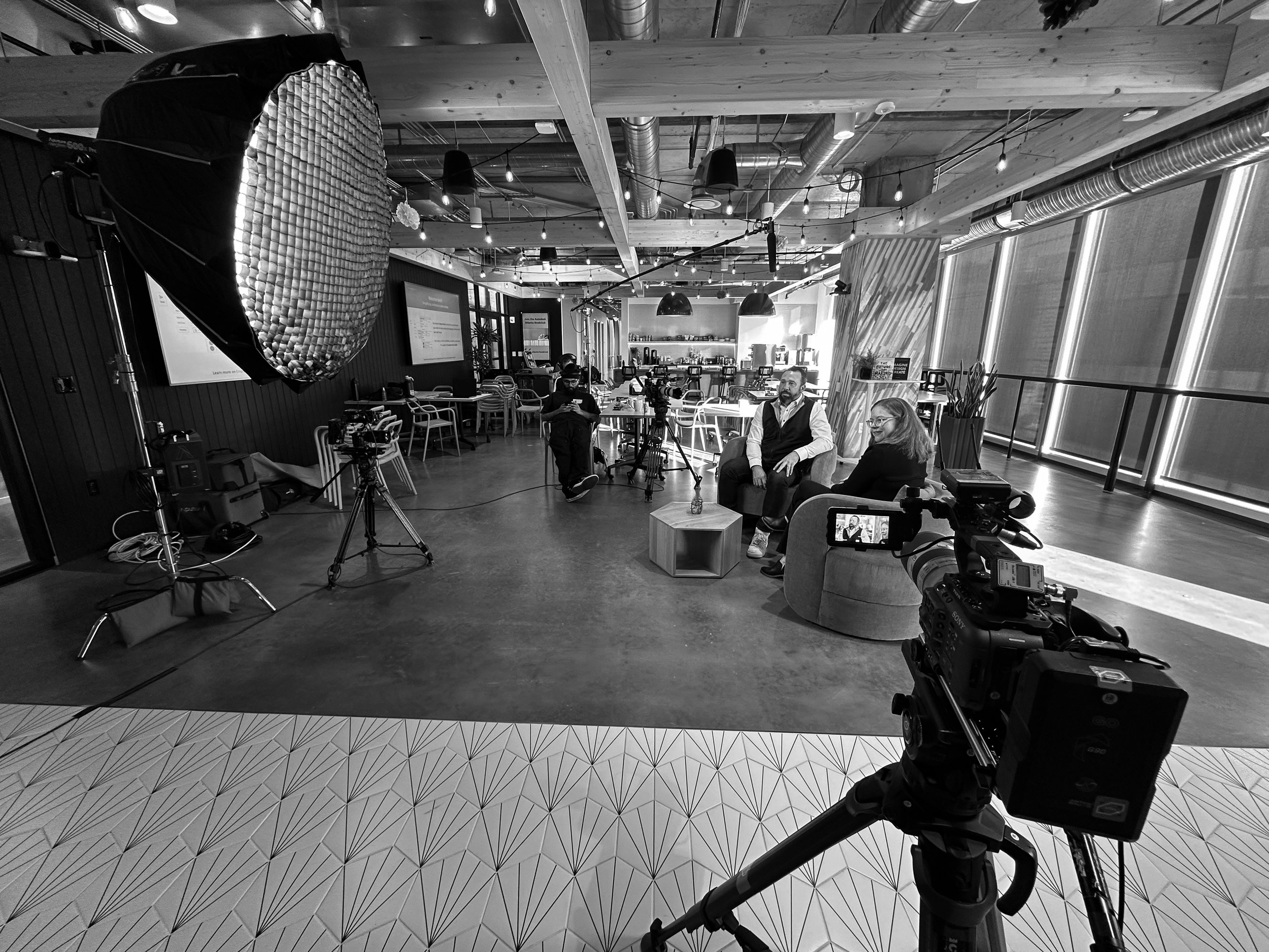 Two people seated in a studio setup being filmed or interviewed, with professional camera equipment, lighting, and a lobby or cafe space in the background.