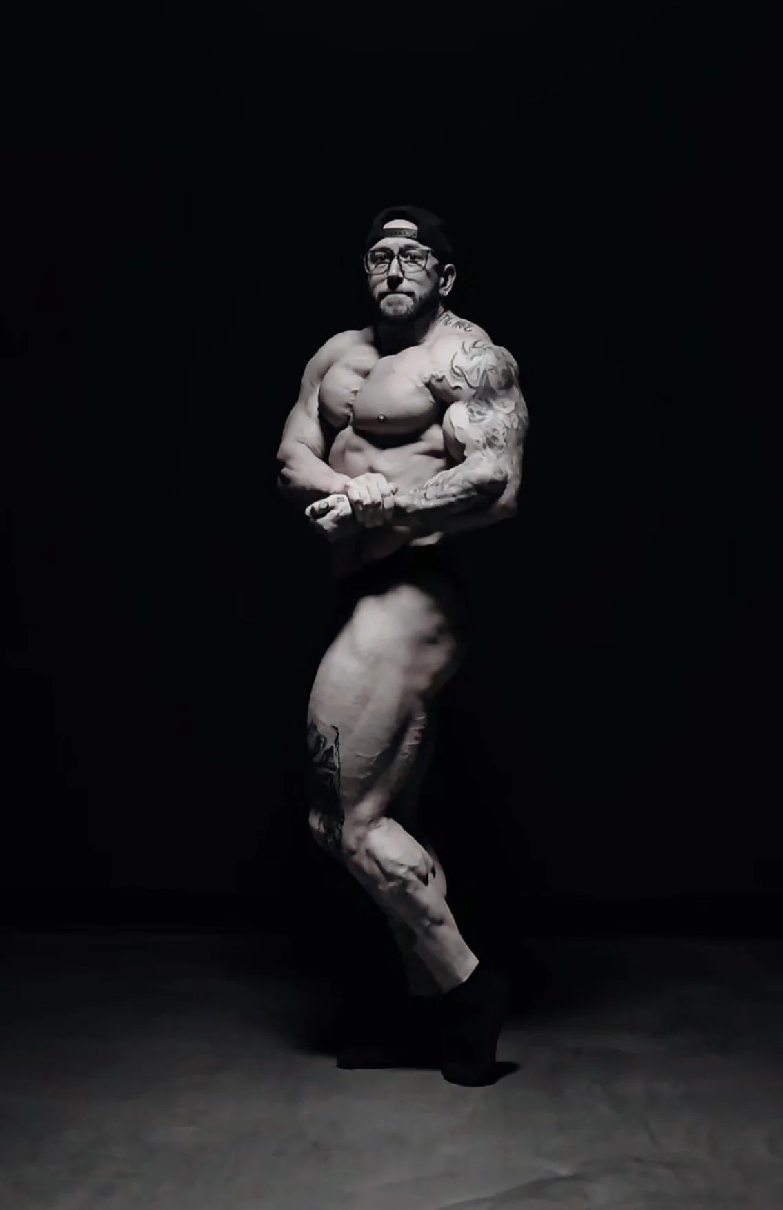 A muscular man with tattoos posing shirtless, wearing a black cap backwards, behind a black background.
