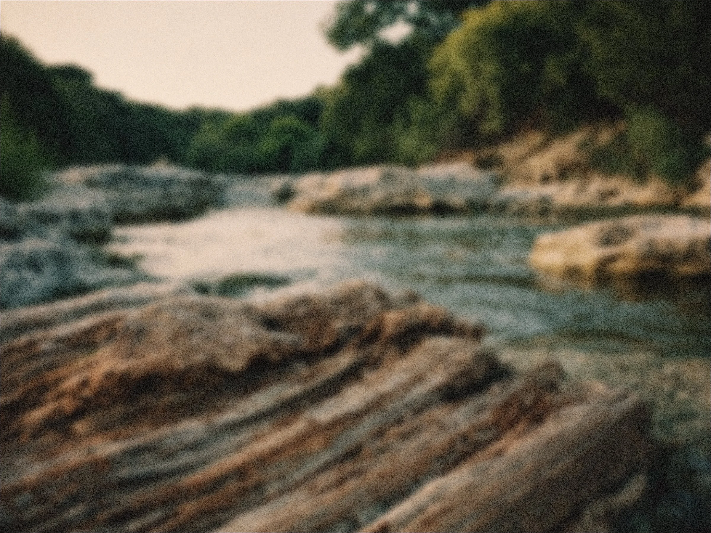 Blurry photo of a river flowing over rocks, surrounded by dense green trees.