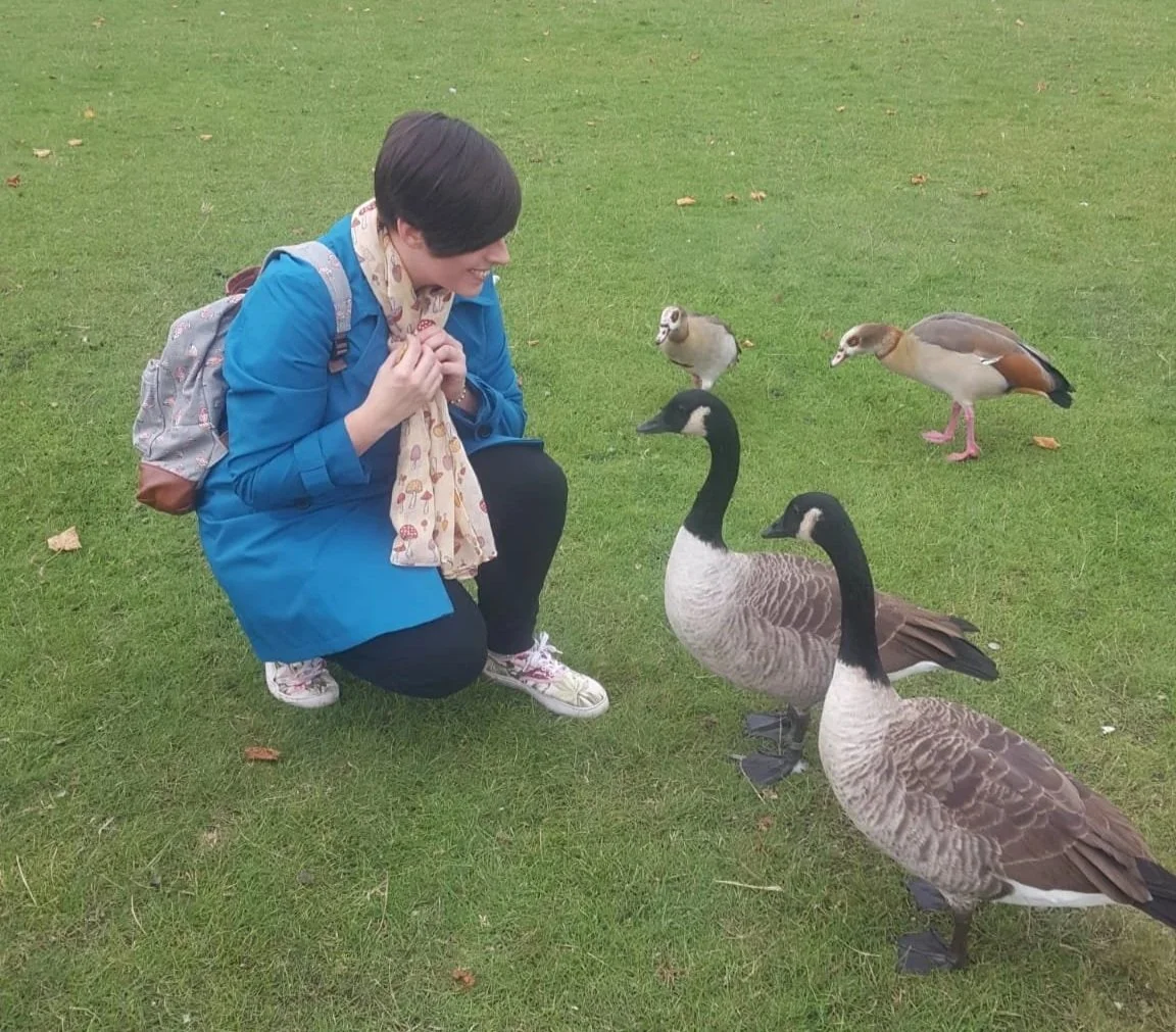 Laura from Tiny Tails Pet Care is crouching on a grassy field, smiling at two Canadian geese nearby, with two more geese in the background.