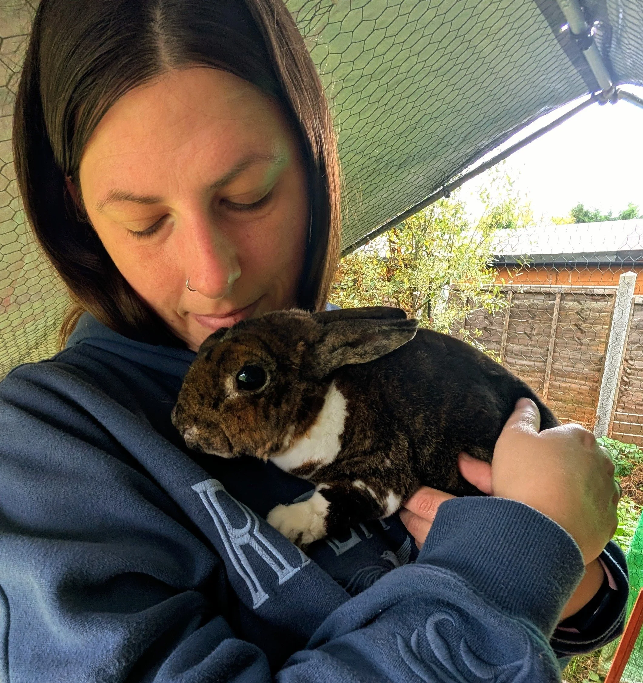 Laura from Tiny Tails Pet Care is holding a brown and black rabbit close to her face, in an outdoor enclosure with a mesh roof and wire fencing.