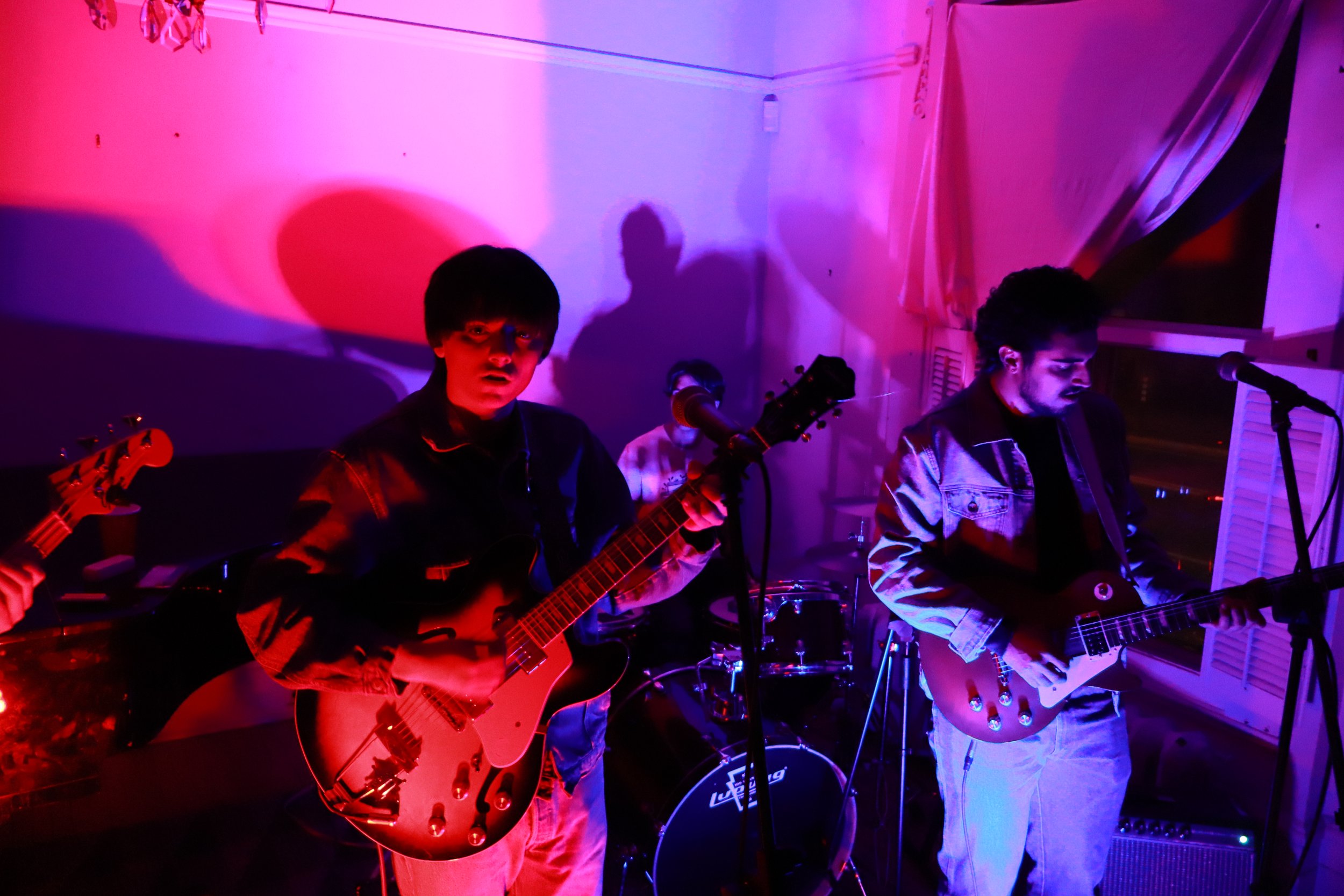 A band performing indoors with colorful purple and red lighting, two guitarists in the foreground, a drummer in the background, and open window with curtains to the right.