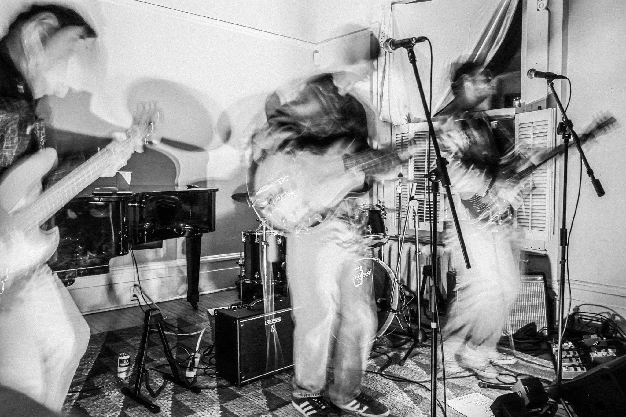 A band of four musicians playing instruments in a room with microphones, music equipment, a drum set, and a grand piano. Motion blur indicates energetic performance.