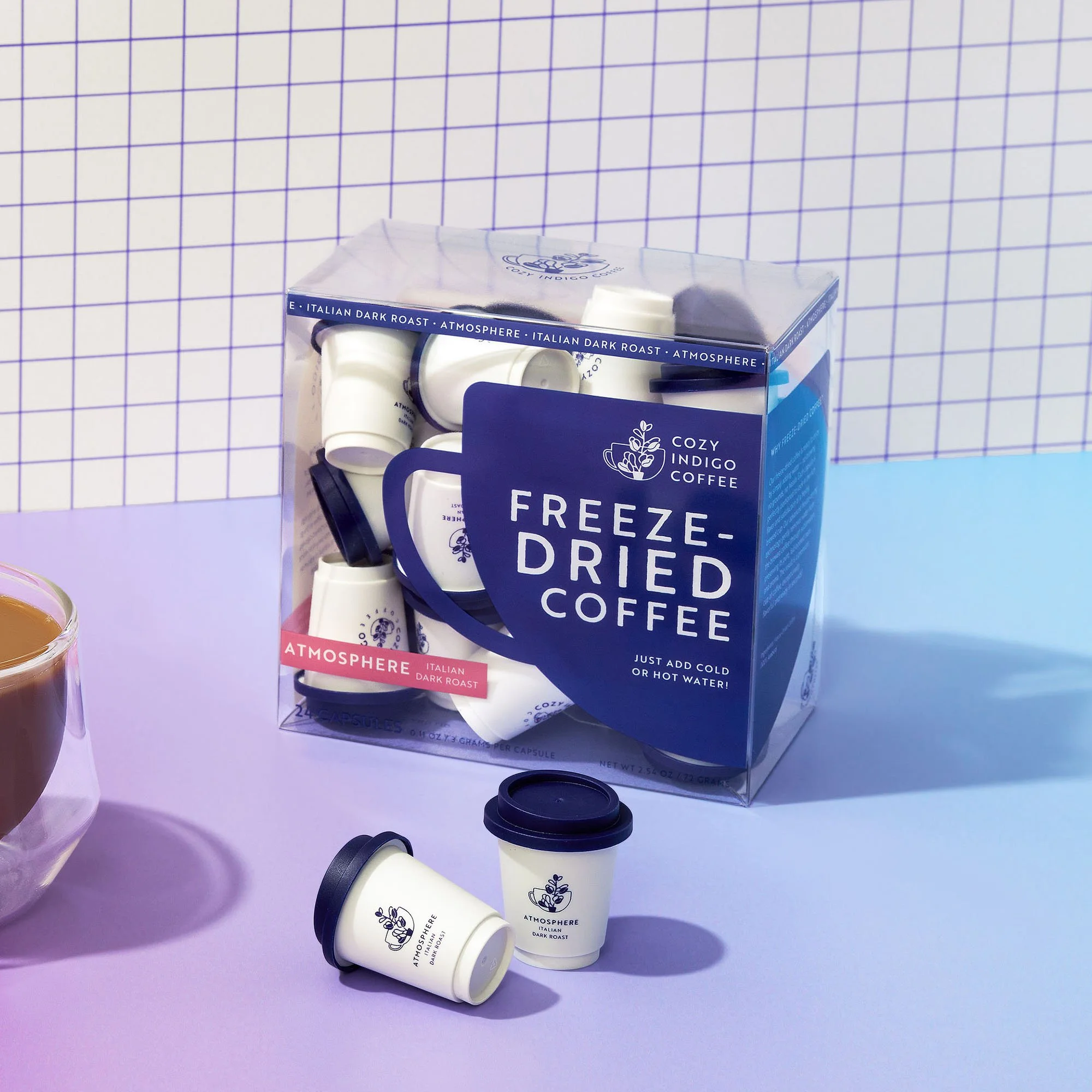A glass cup filled with coffee and two small coffee cups with dark blue lids, next to a box of ATMOSPHERE freeze-dried coffee capsules on a purple surface with a purple and white grid background.