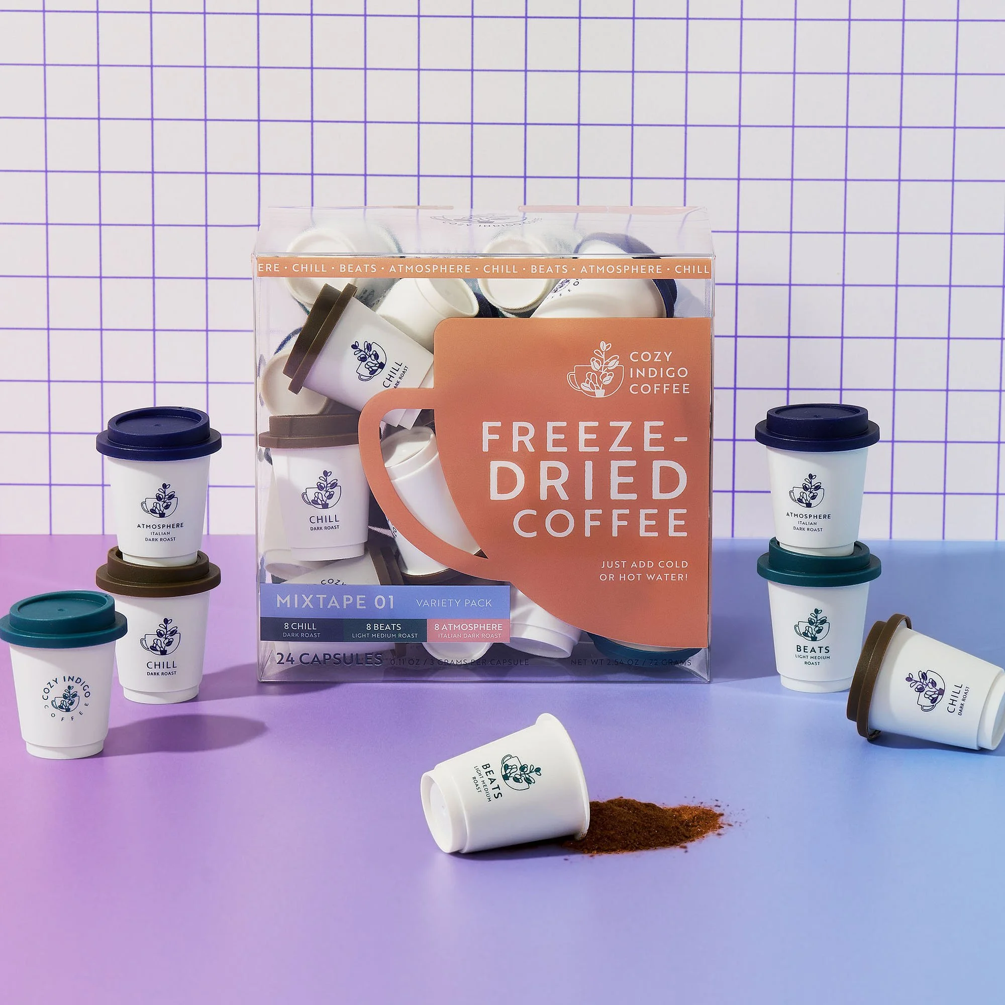 A box of MIXTAPE 01 freeze-dried Coffee capsules with colorful cups and a spilled coffee grounds on a purple surface, with an indigo and cream grid background.