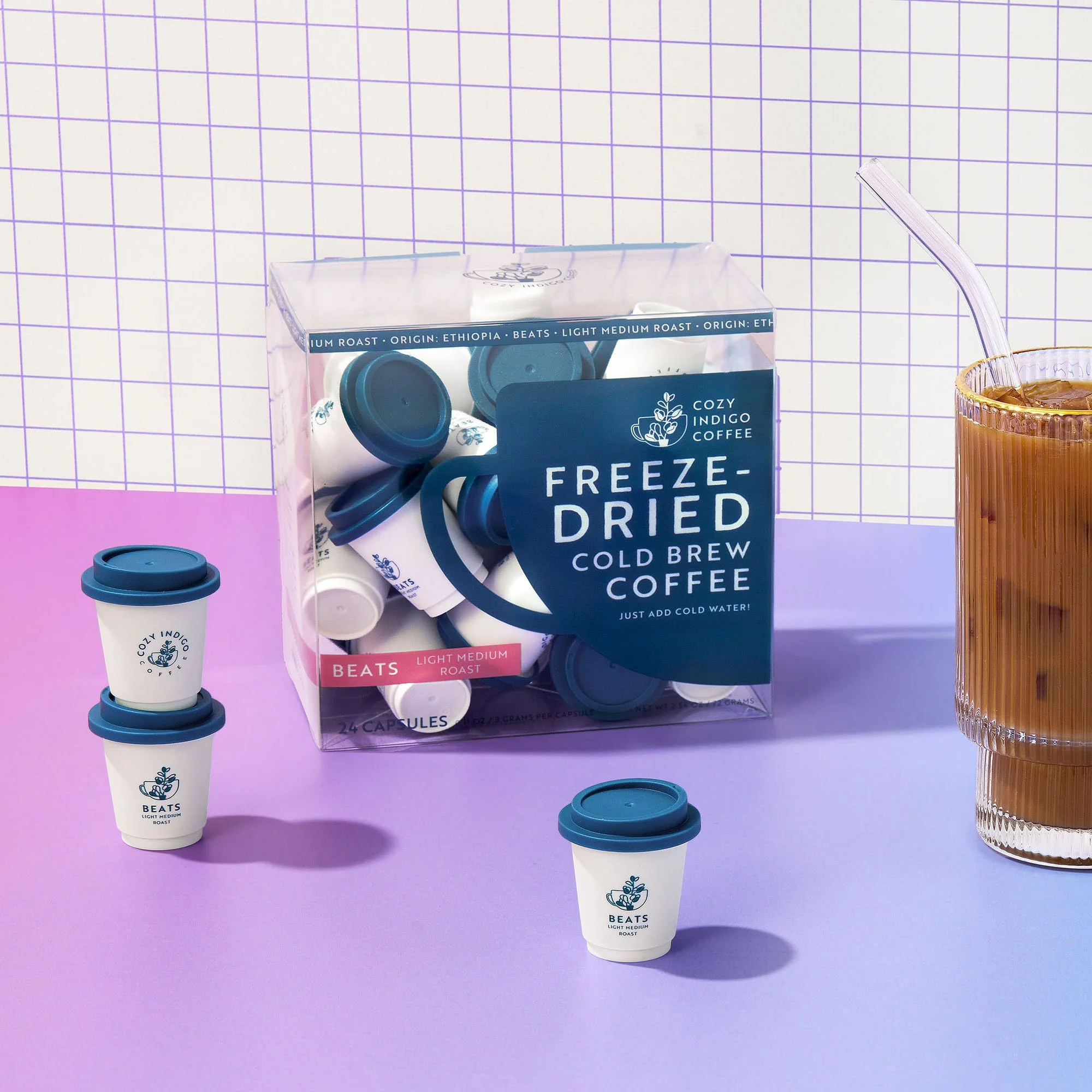 Box of BEATS freeze-dried cold brew coffee capsules next to a glass of iced coffee with a straw on a purple surface with a white and purple grid background.