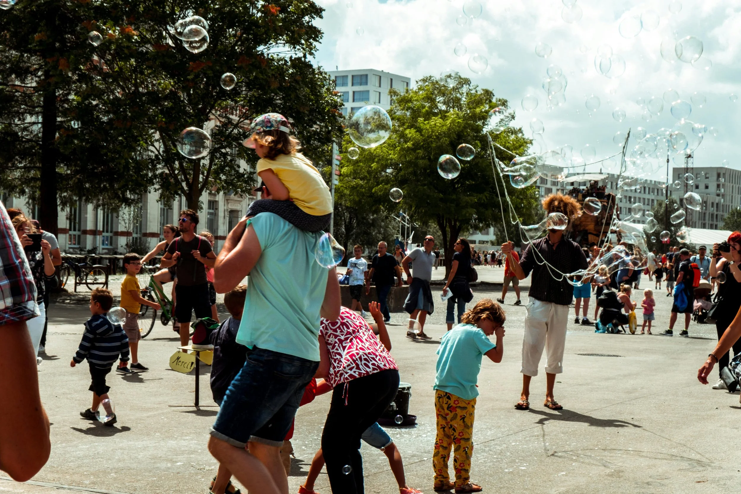 A lively outdoor scene with people of various ages enjoying a street festival, including children playing with bubbles, a performer creating bubbles, and families strolling. Trees and modern buildings are visible in the background.