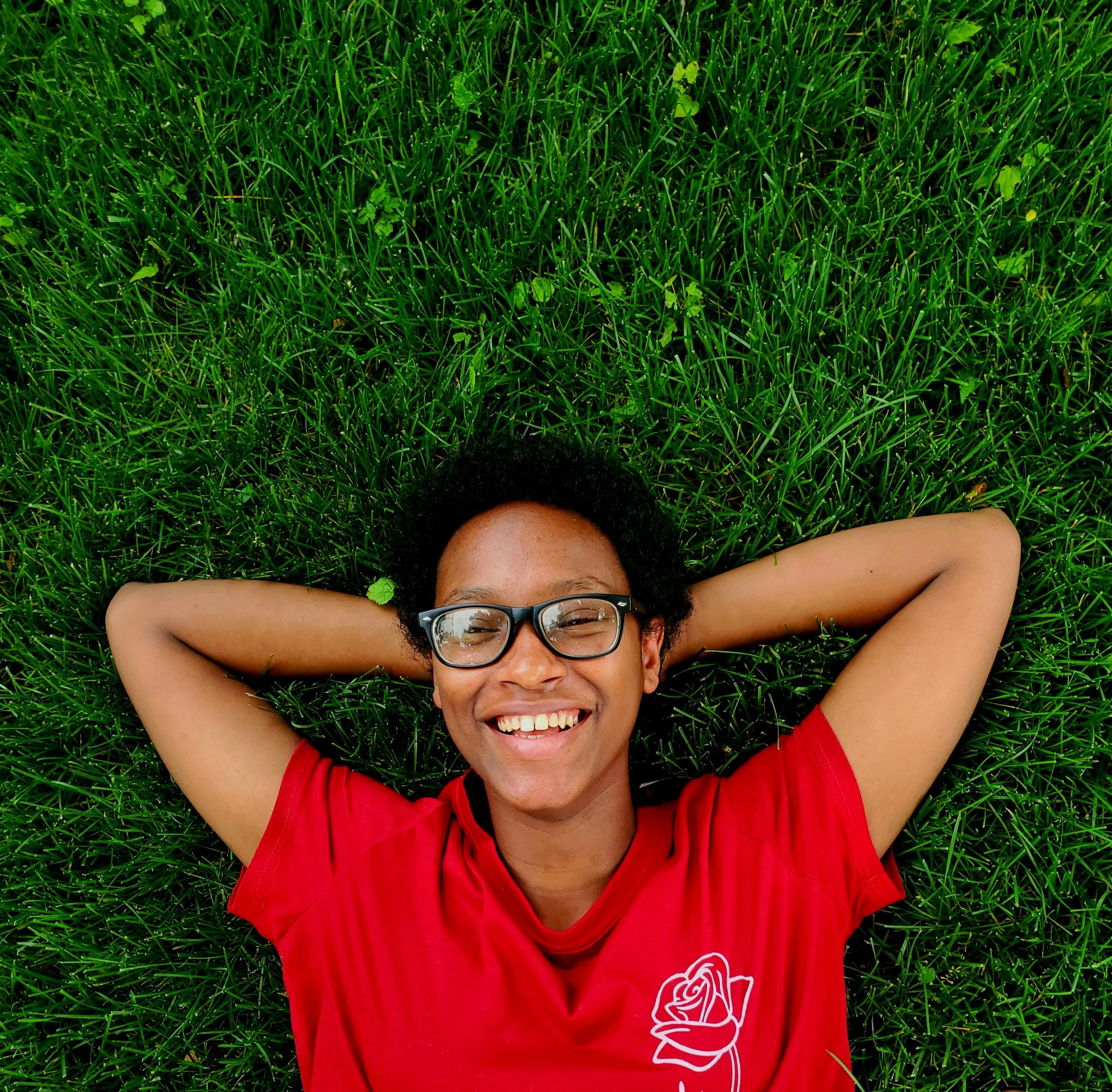 A smiling person with short curly black hair, glasses, and a red shirt with a white rose design, lying on green grass with arms behind their head.