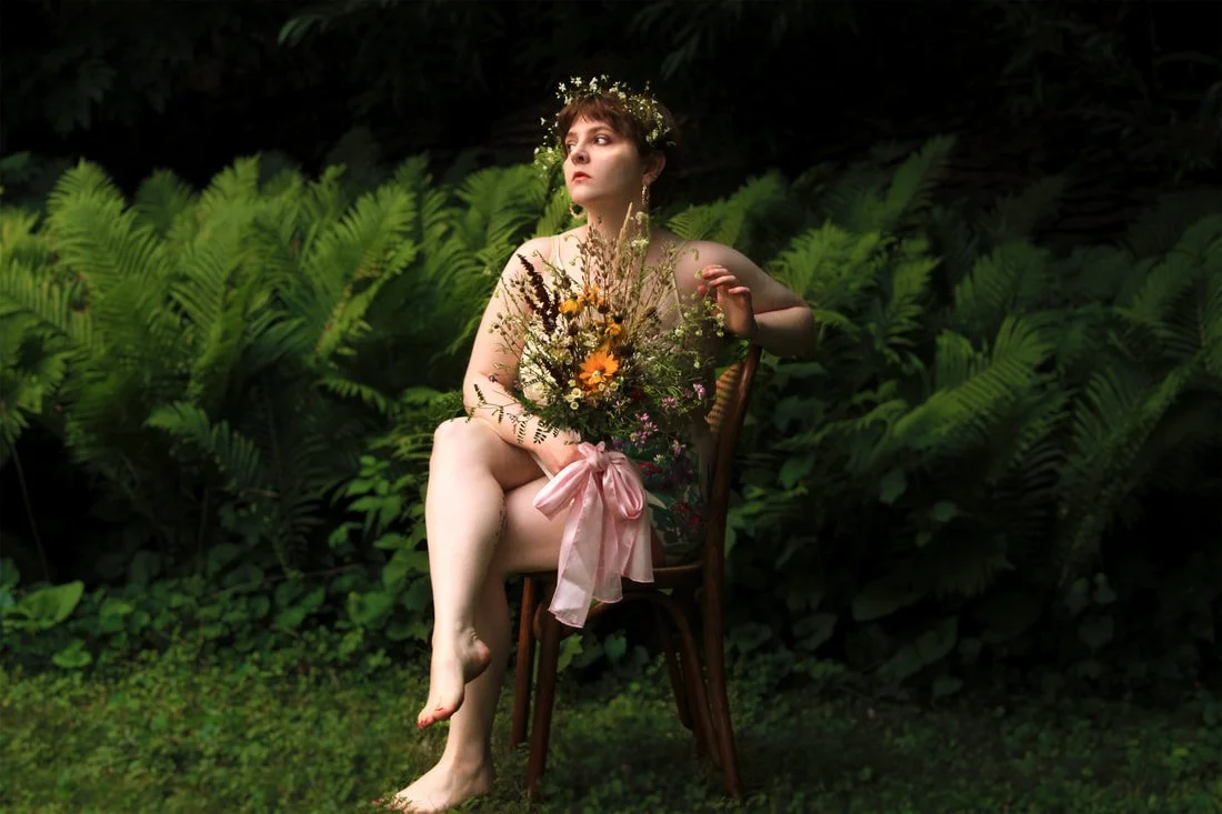 A woman with short brown hair and a floral crown sits on a wooden chair amid lush green ferns, holding a bouquet of flowers wrapped with a pink ribbon, with a serene expression.