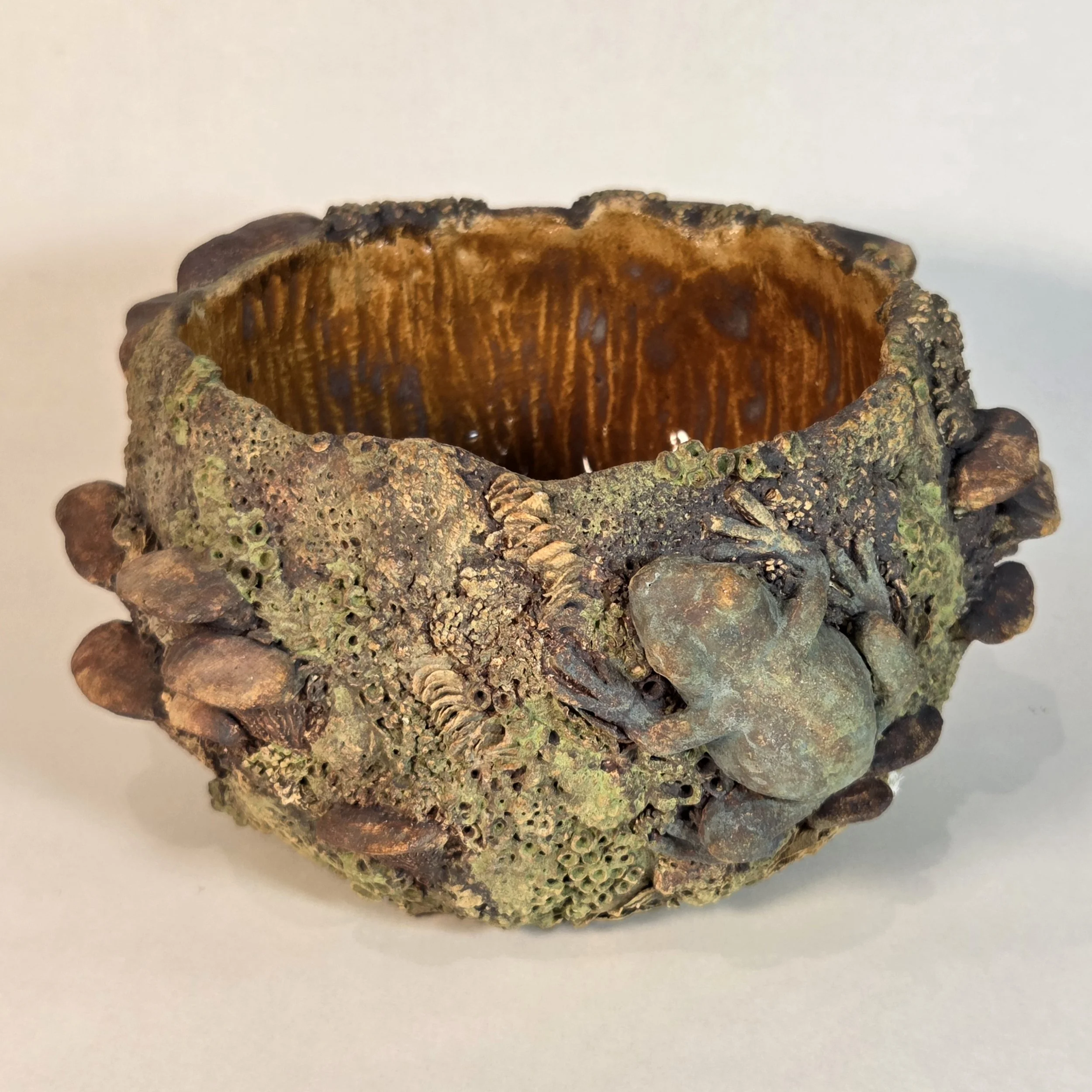 A decorative bowl made from natural materials, featuring a frog figurine on the side, with a textured, moss-like appearance and an open interior.