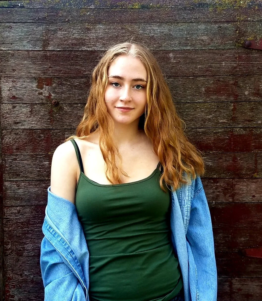 A young woman with wavy red hair, wearing a green tank top and a blue denim jacket, standing in front of a weathered wooden wall.