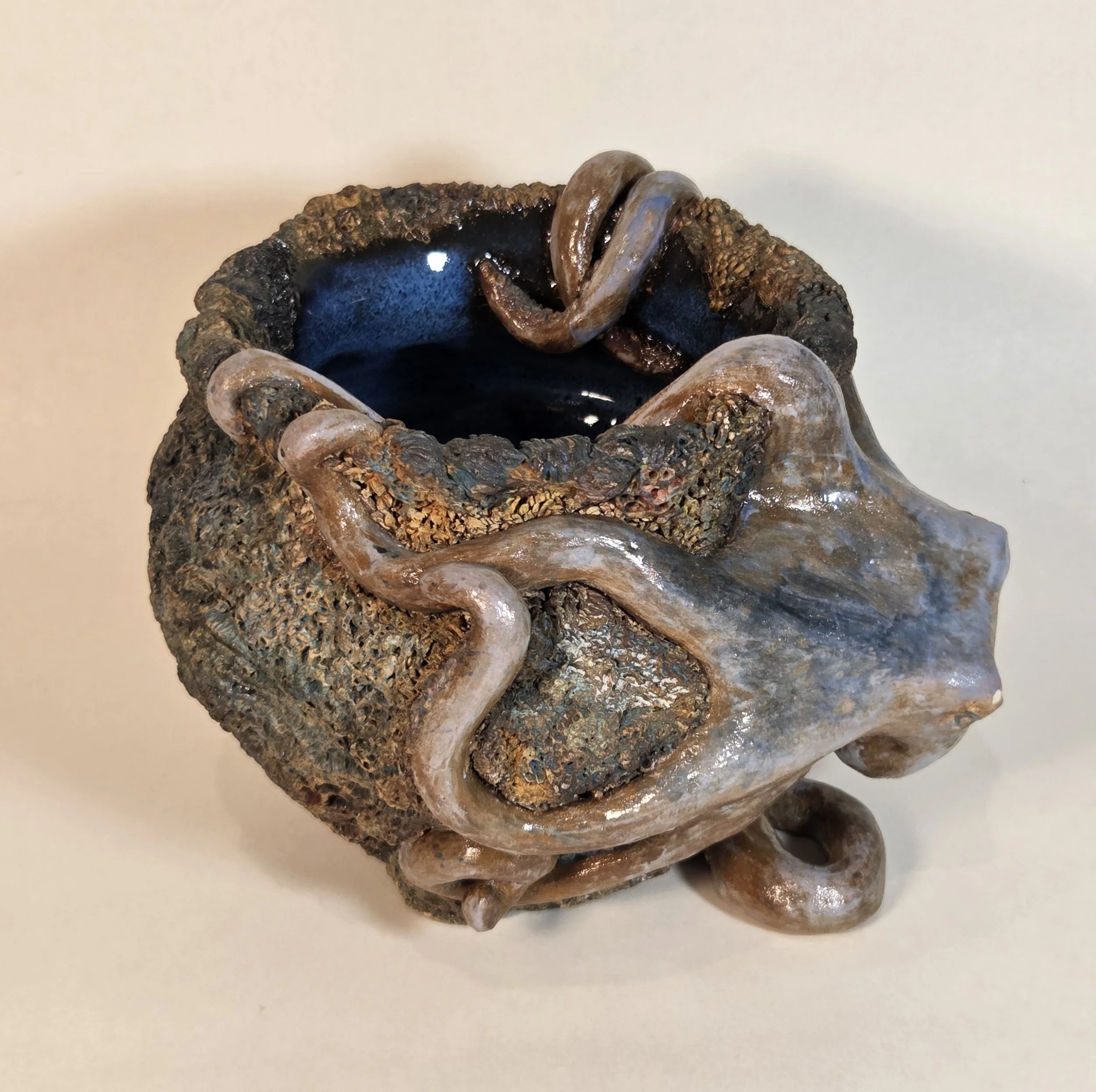 A ceramic vessel with a textured, dark exterior and a smooth, light-colored face design, decorated with serpentine figures wrapping around the surface.