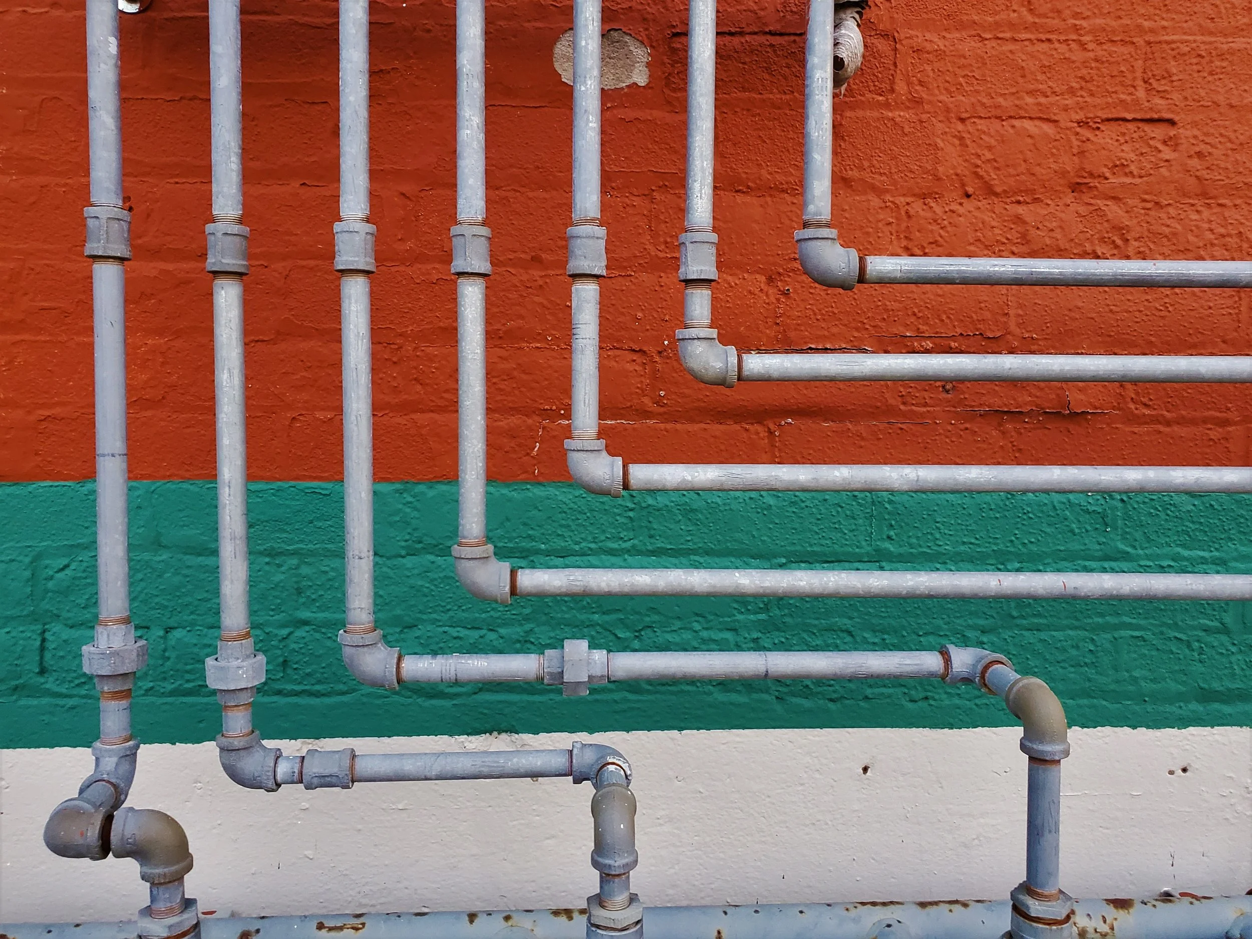 Metal pipes installed against a wall painted in three horizontal stripes: orange at the top, green in the middle, and white at the bottom.