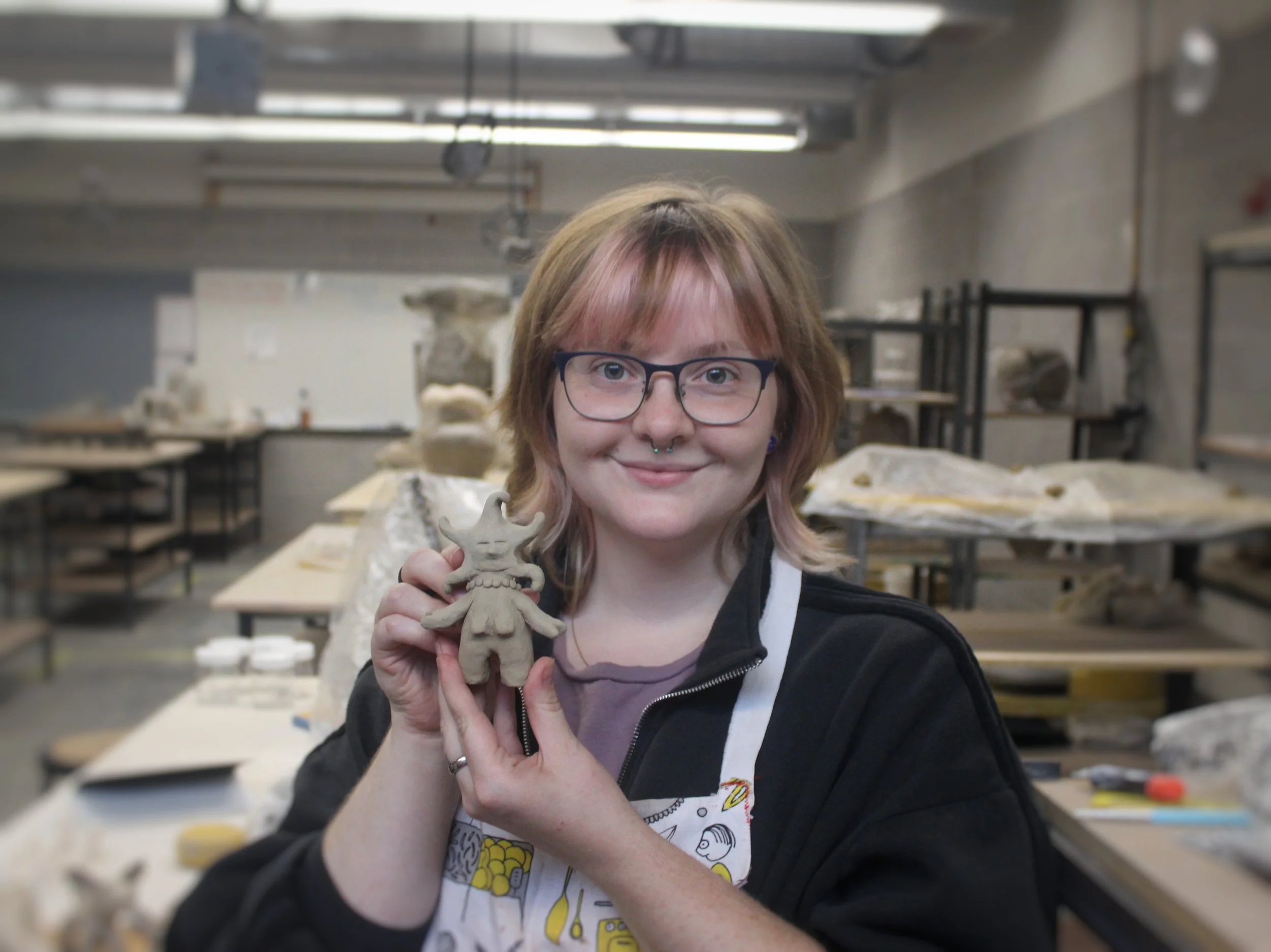 A woman with glasses and pinkish-brown hair holding a small, detailed clay figurine in a ceramic studio.