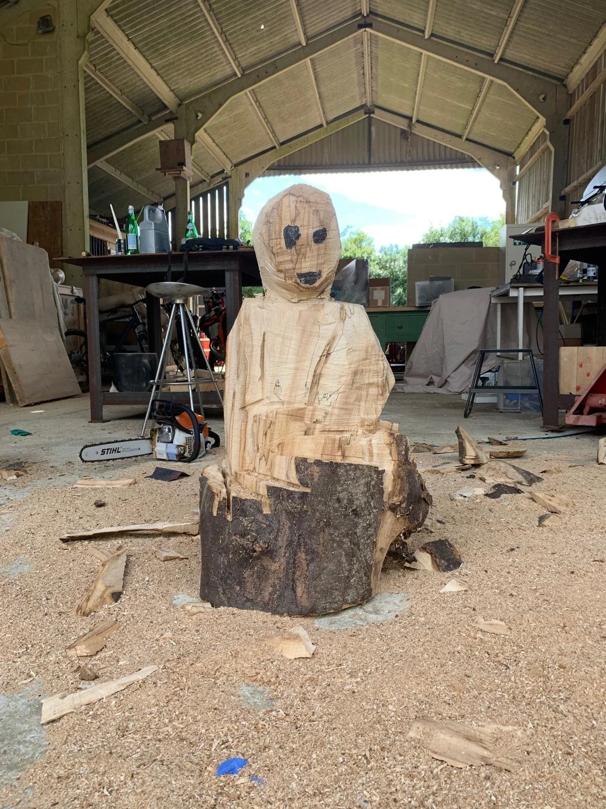A wooden sculpture of a person with a simple face carved into the wood, sitting on a tree stump inside a woodworking workshop.