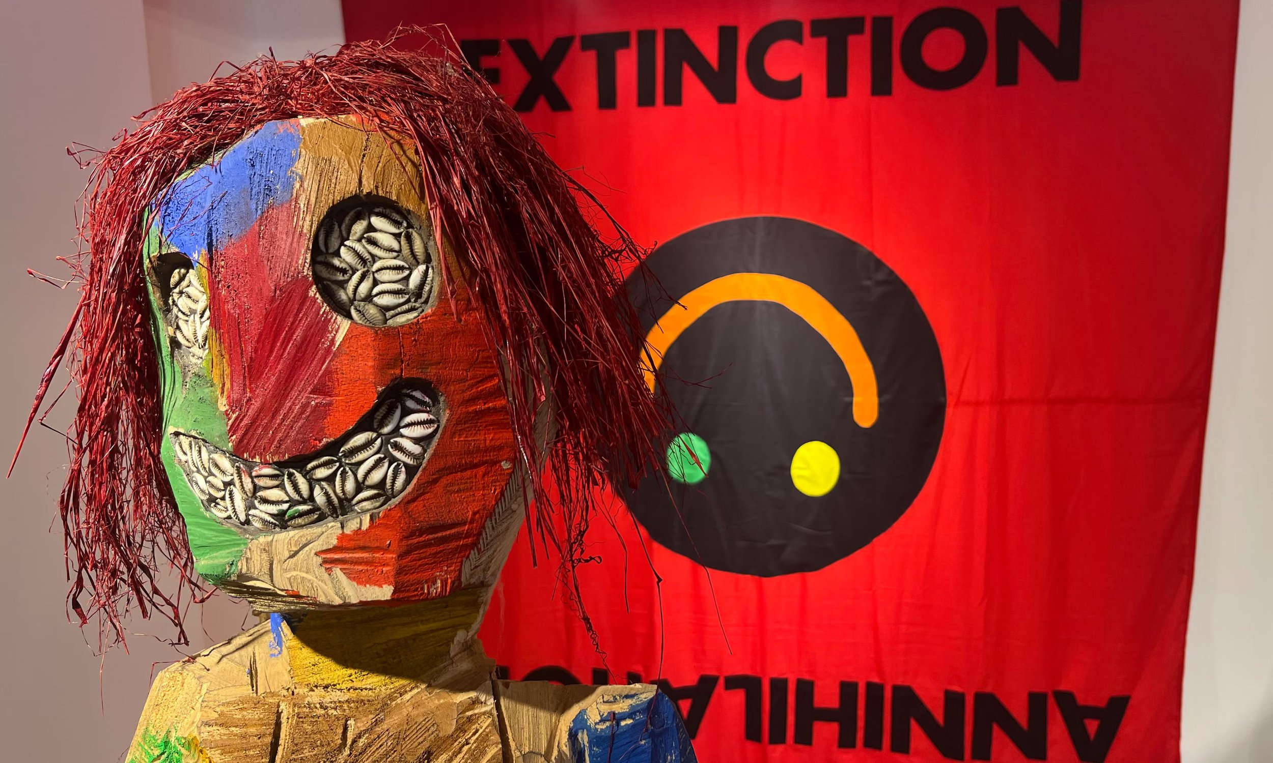 A colorful, hand-painted wooden puppet with red, blue, green, and yellow patches, featuring a face with a large smile made of sunflower seeds, red textured hair made of thin fibers, and a painted body. In the background, there is a red banner with th