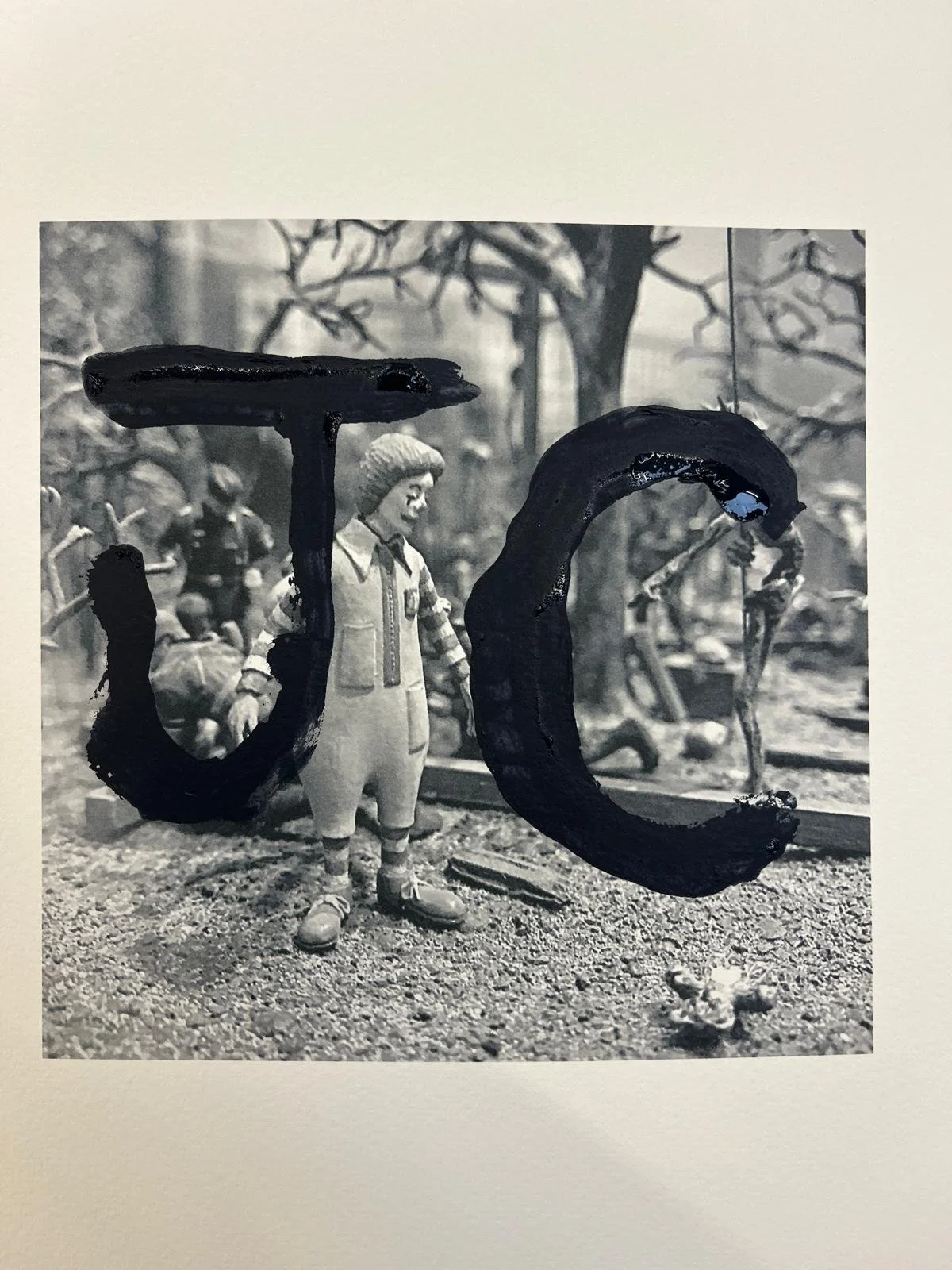 A black and white photograph with painted black letters spelling 'TOC' in front of a toy soldier figure, a large tree in the background, and other toy soldiers scattered on the ground.