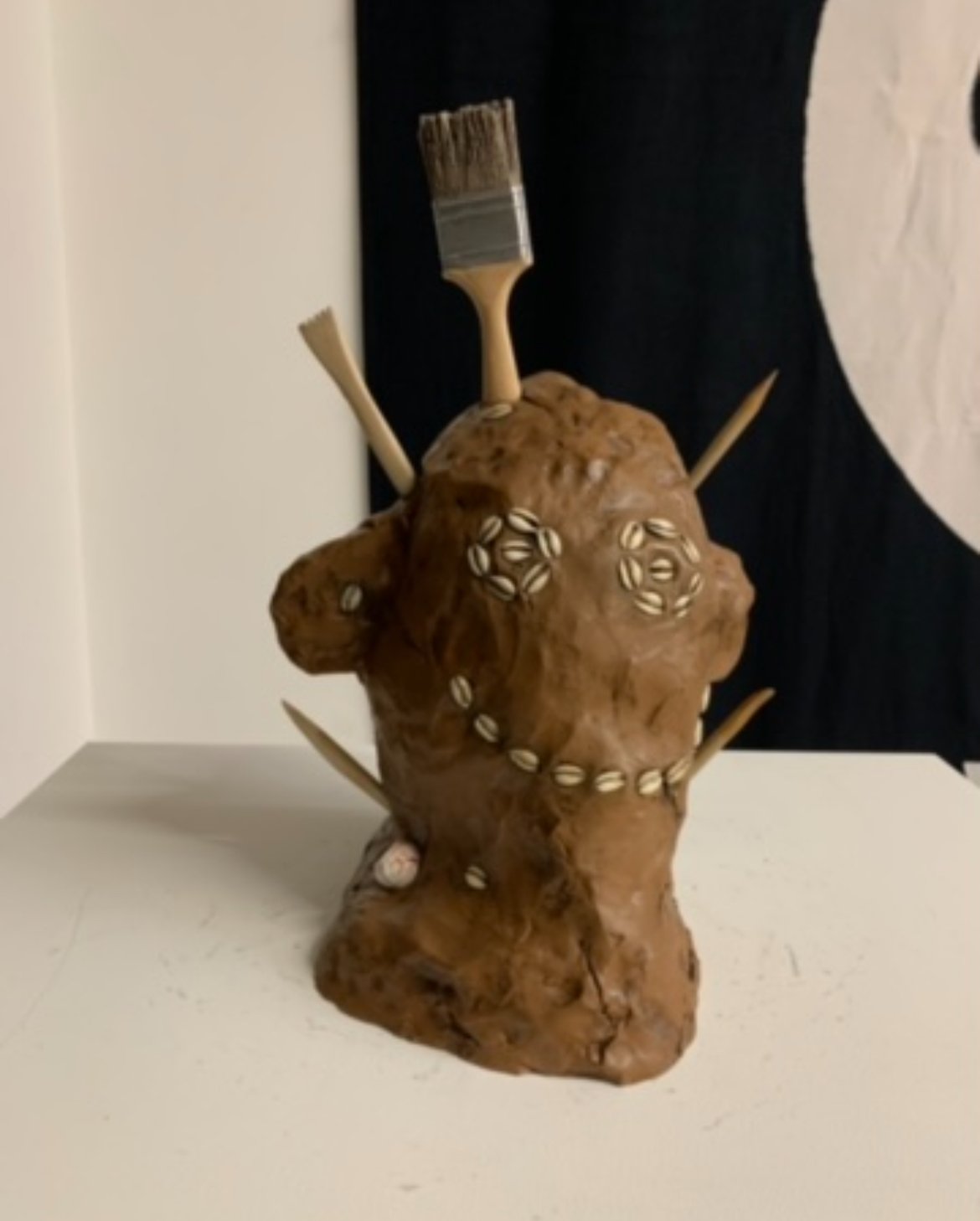 Decorative sculpture resembling a face made of a large, brown, textured rock with shells embedded, with small brushes and sticks protruding from it.