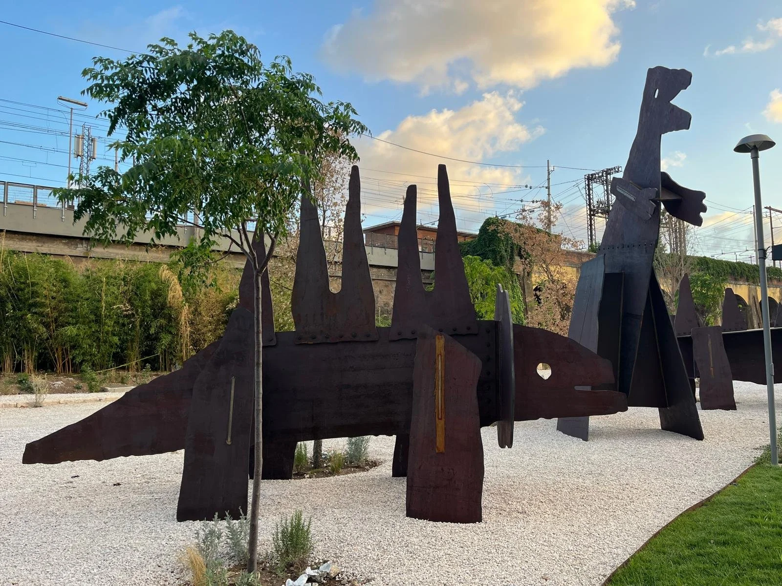 Outdoor sculpture resembling a dinosaur made of large, dark metal pieces with a long neck and head, surrounded by trees and a gravel ground.