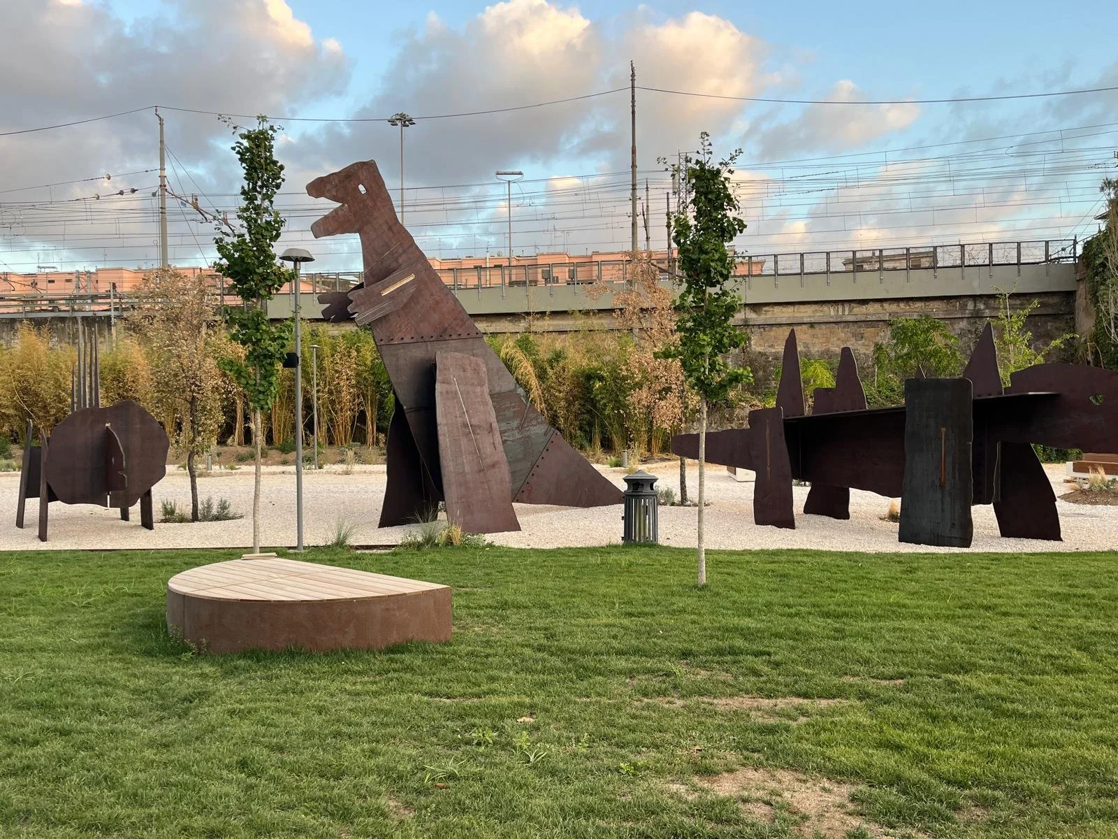 Outdoor park with large rust-colored metal sculptures, including a tall dinosaur and abstract shapes, surrounded by green grass and small trees, under a partly cloudy sky.