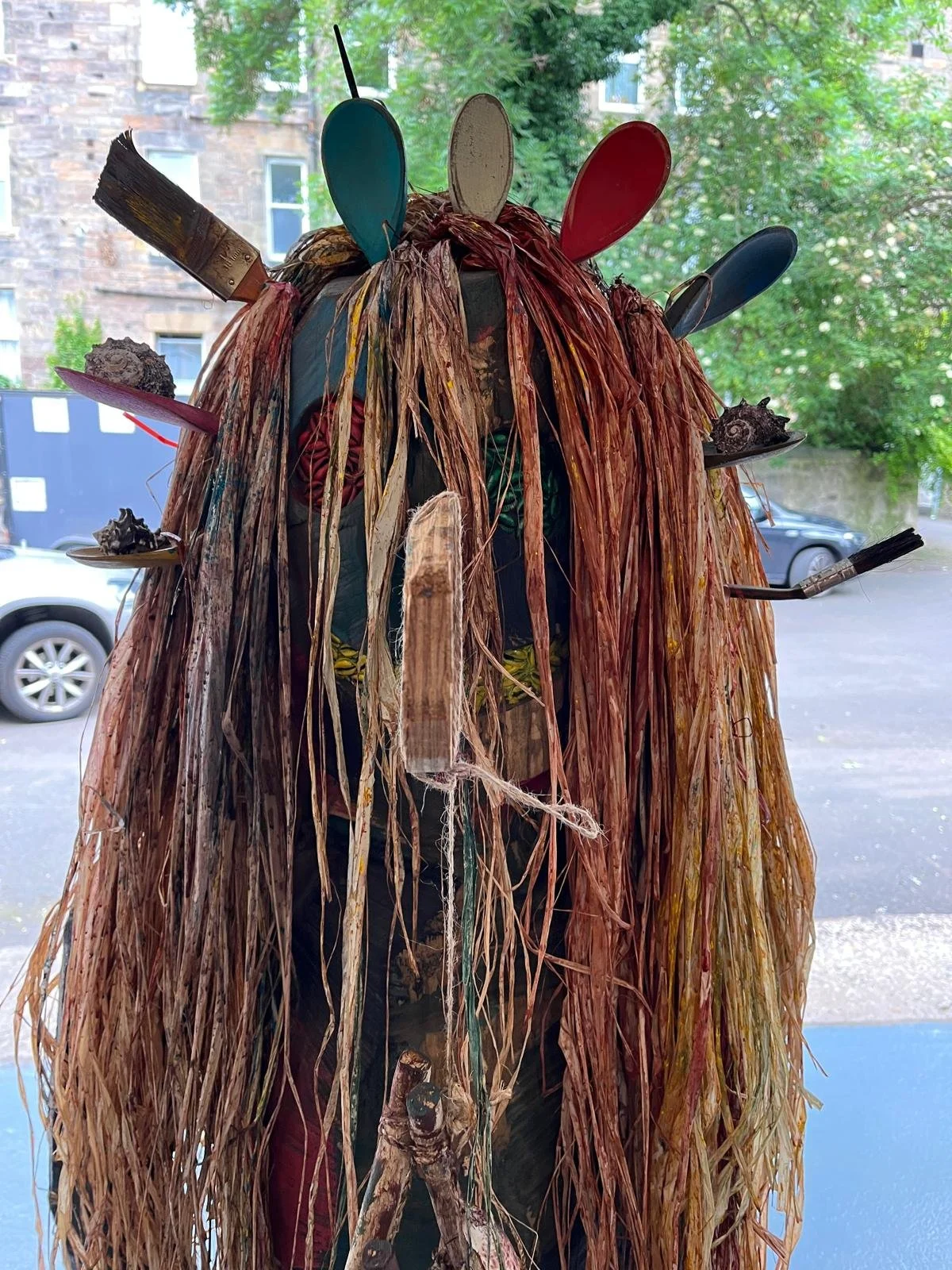 A costume with a long, shaggy, reddish-brown fiber wig adorned with colorful spoons, paintbrushes, and shells.